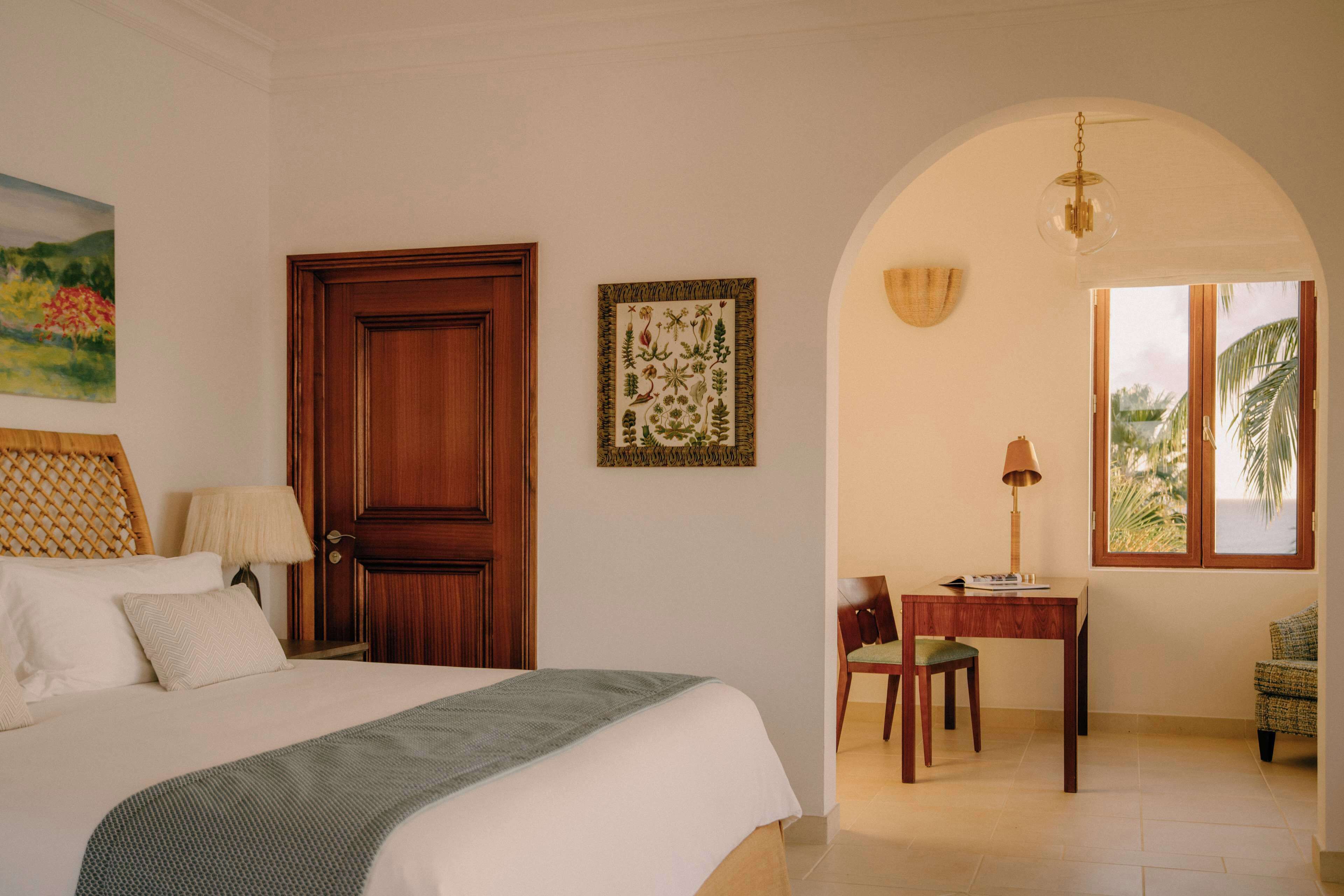 A bedroom with a bed, a wooden door, a framed botanical illustration, a desk with a chair, and a window with a view of palm trees.