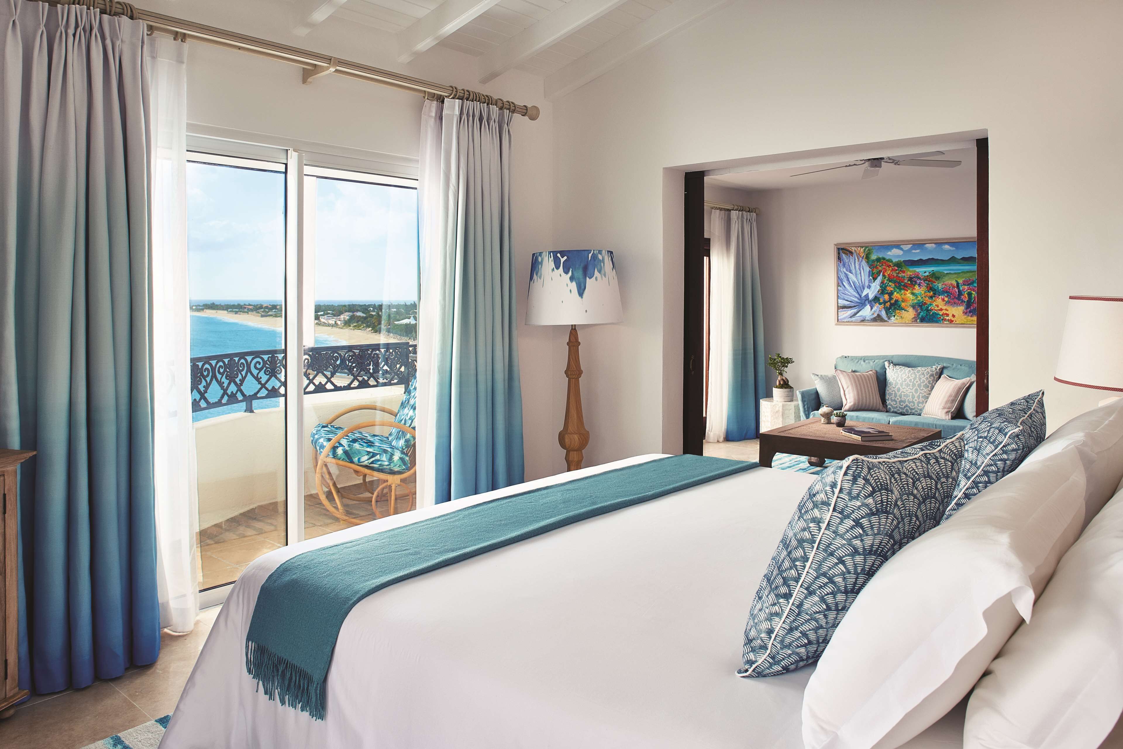 A hotel room with a large bed, a balcony with a view of the beach, a painting on the wall, and a chair by the window.