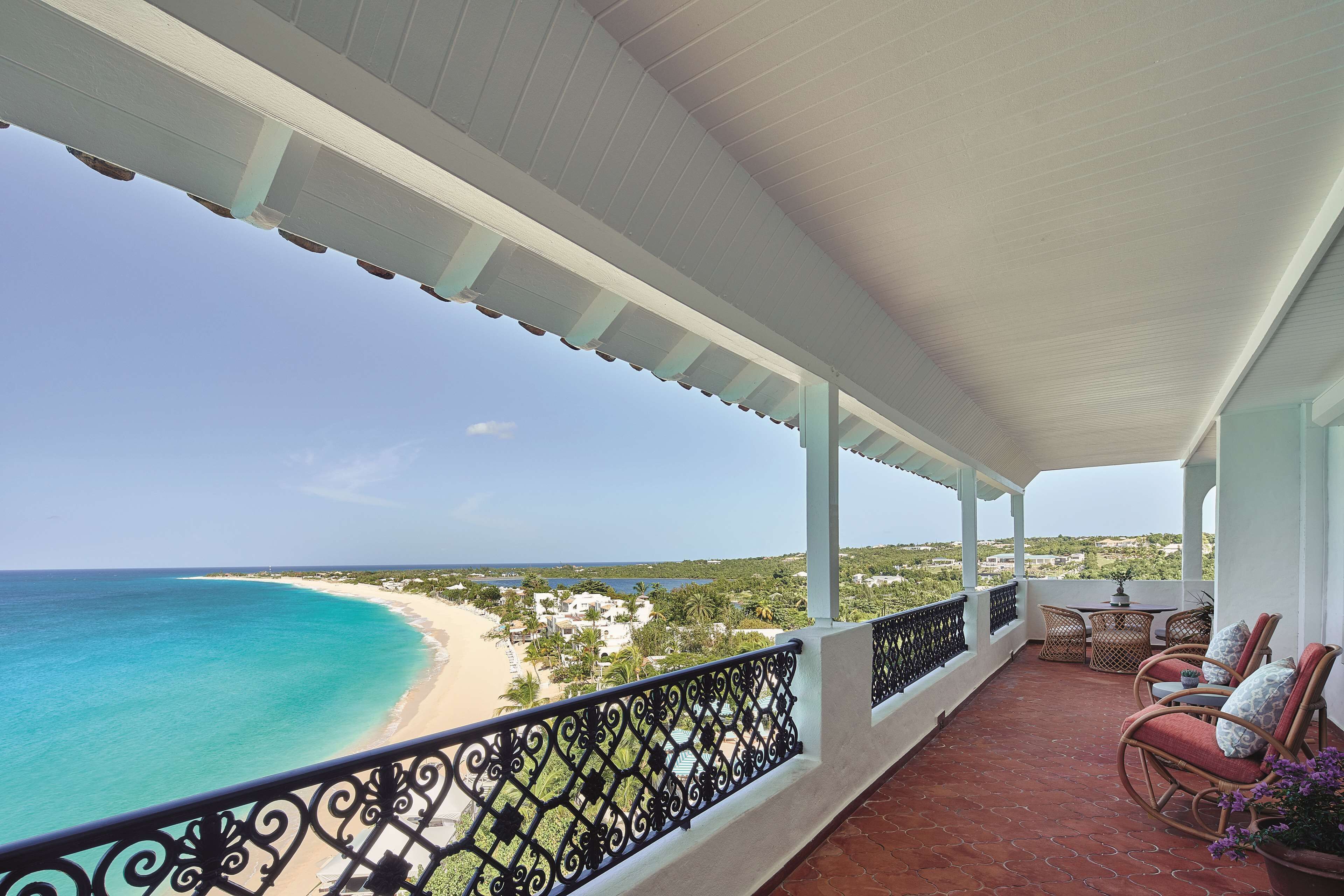 A balcony with a view of a beach and ocean, wicker chairs, and a table.
