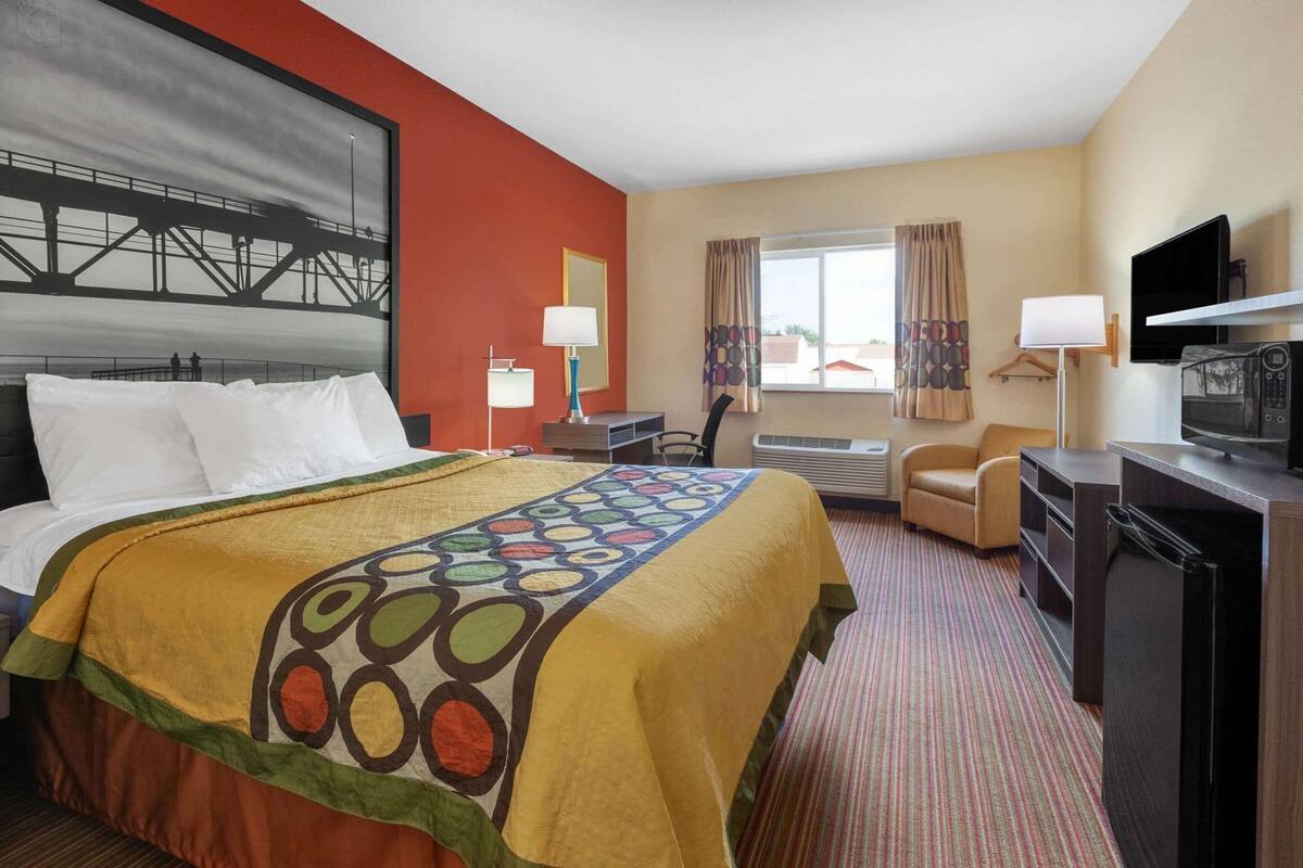 Quarto de hotel com cama, escrivaninha, cadeira, televisão e janela com cortinas.