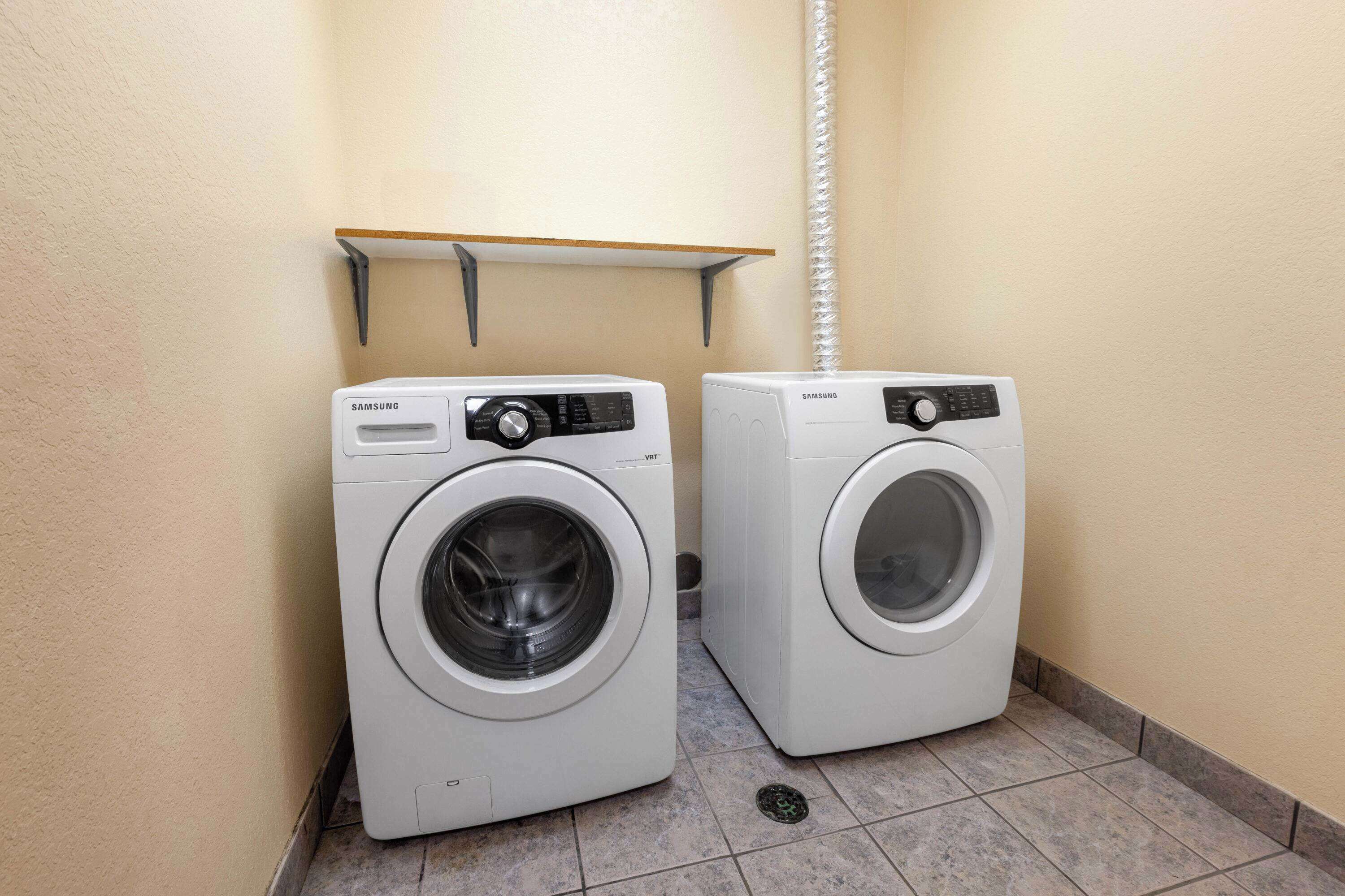 Two white Samsung front-loading washing machines in a corner with a wooden shelf above them.