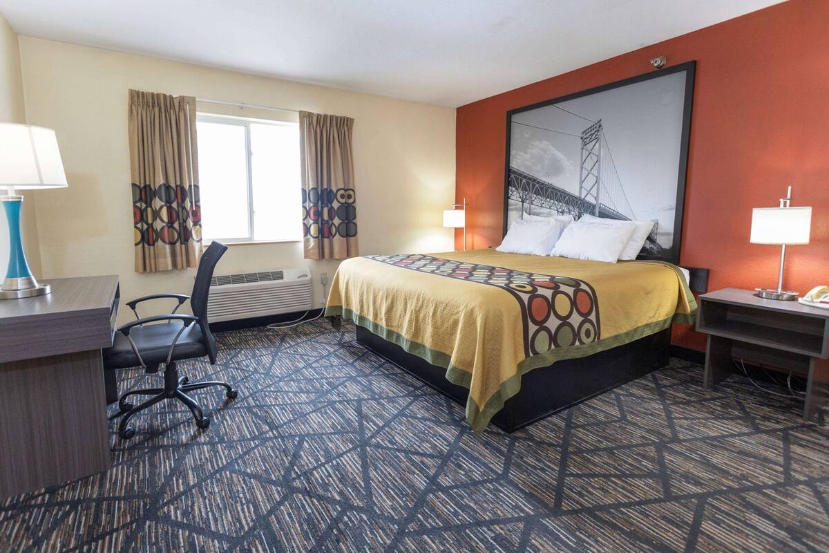 Quarto de hotel com uma cama grande, uma escrivaninha, uma cadeira, uma janela com cortinas e um quadro na parede.
