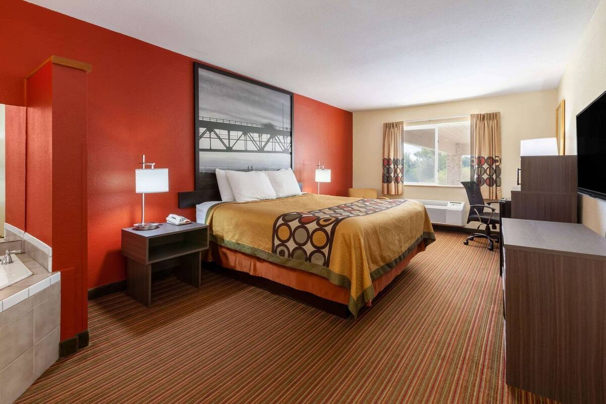 Quarto de hotel com cama, mesas de cabeceira, escrivaninha, cadeira, televisão e janela com cortinas.