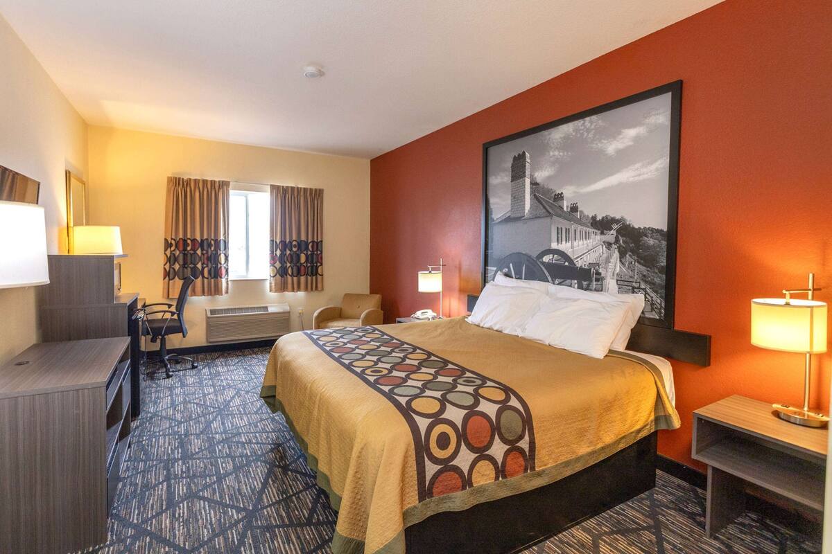Quarto de hotel com uma cama grande, uma escrivaninha, uma cadeira, abajur e uma moldura grande na parede.