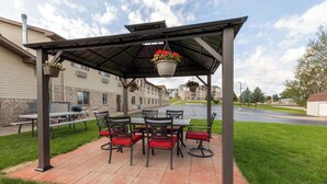 Un patio cubierto con mesa y sillas, un macramé colgante con flores y un sendero de ladrillos.
