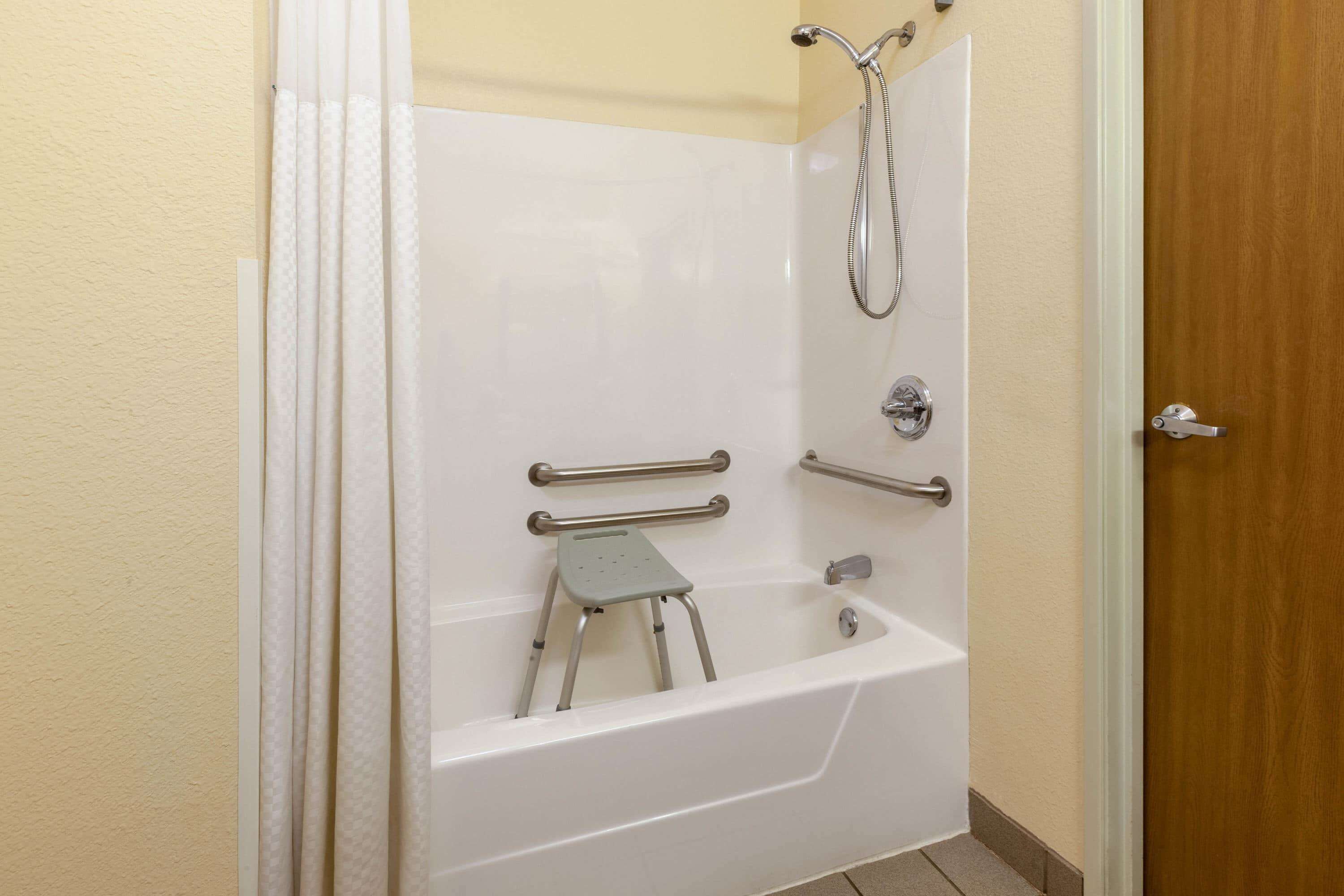 Un baño con una bañera blanca, barras de apoyo, un cabezal de ducha y un pequeño taburete.
