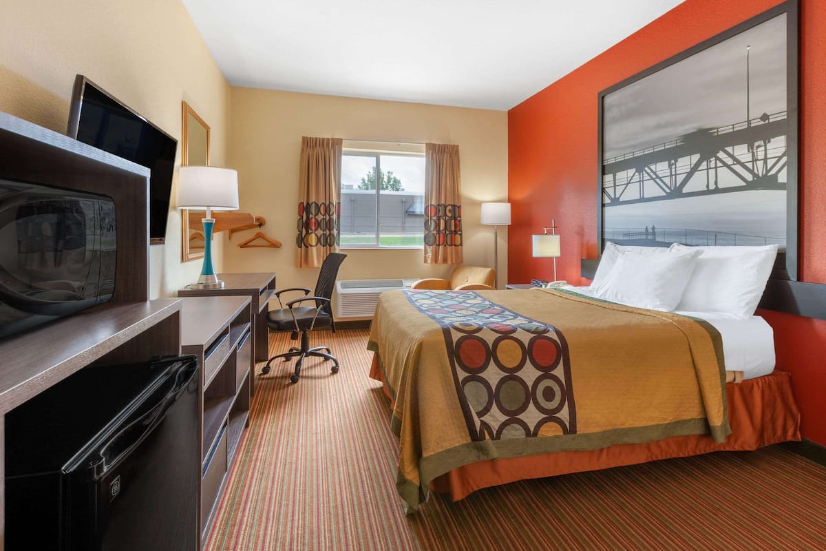 Quarto de hotel com cama, escrivaninha, televisão e uma janela grande com cortinas.