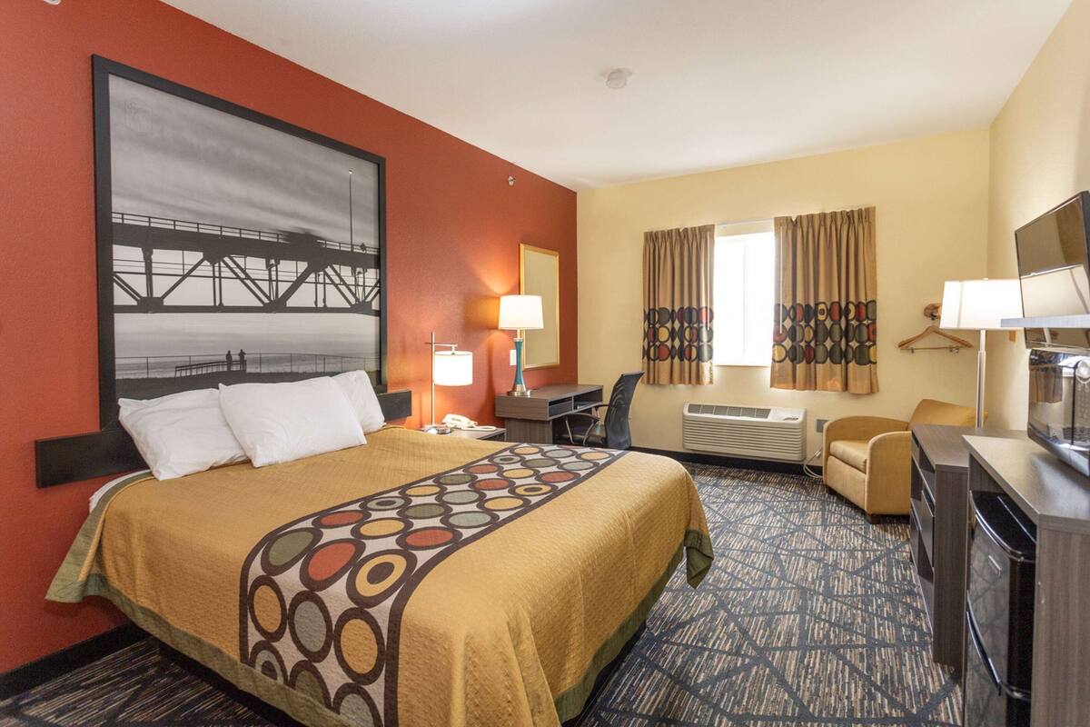 Quarto de hotel com cama, escrivaninha, cadeira, televisão e um quadro grande na parede.