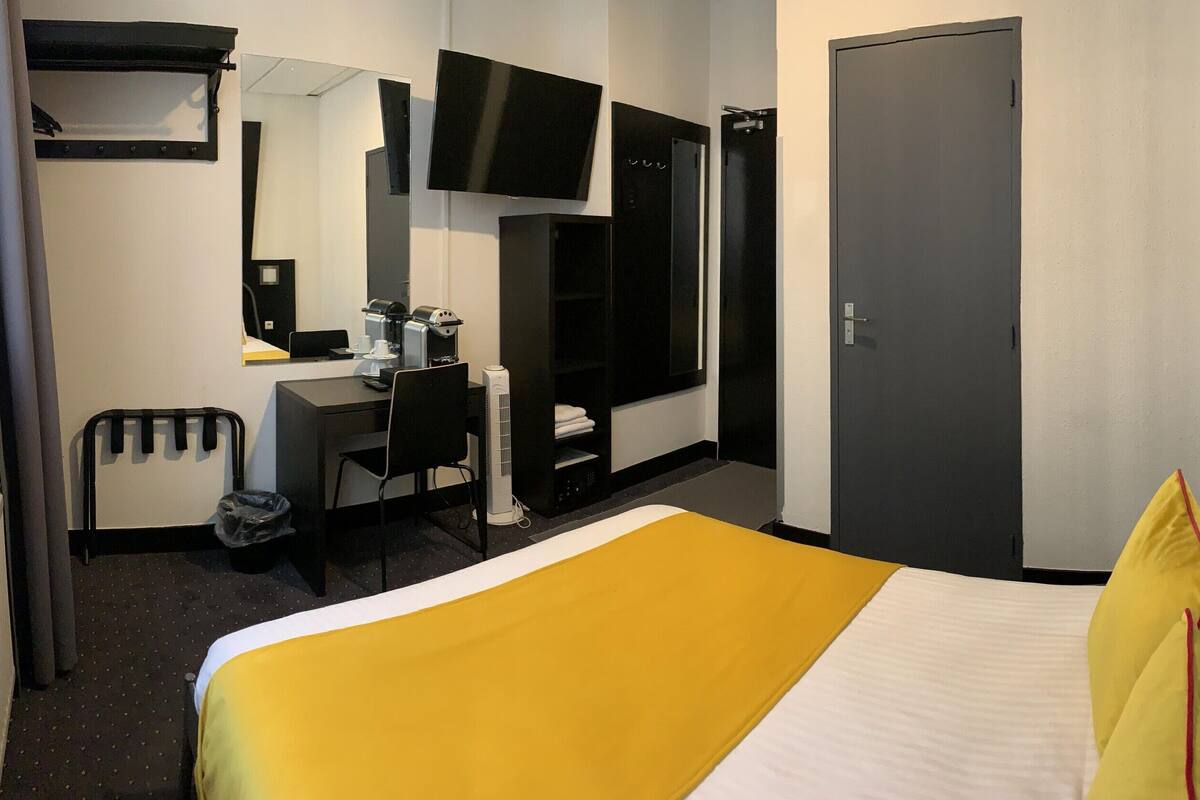 Habitación de hotel con cama, escritorio, silla, cafetera, espejo y televisión.