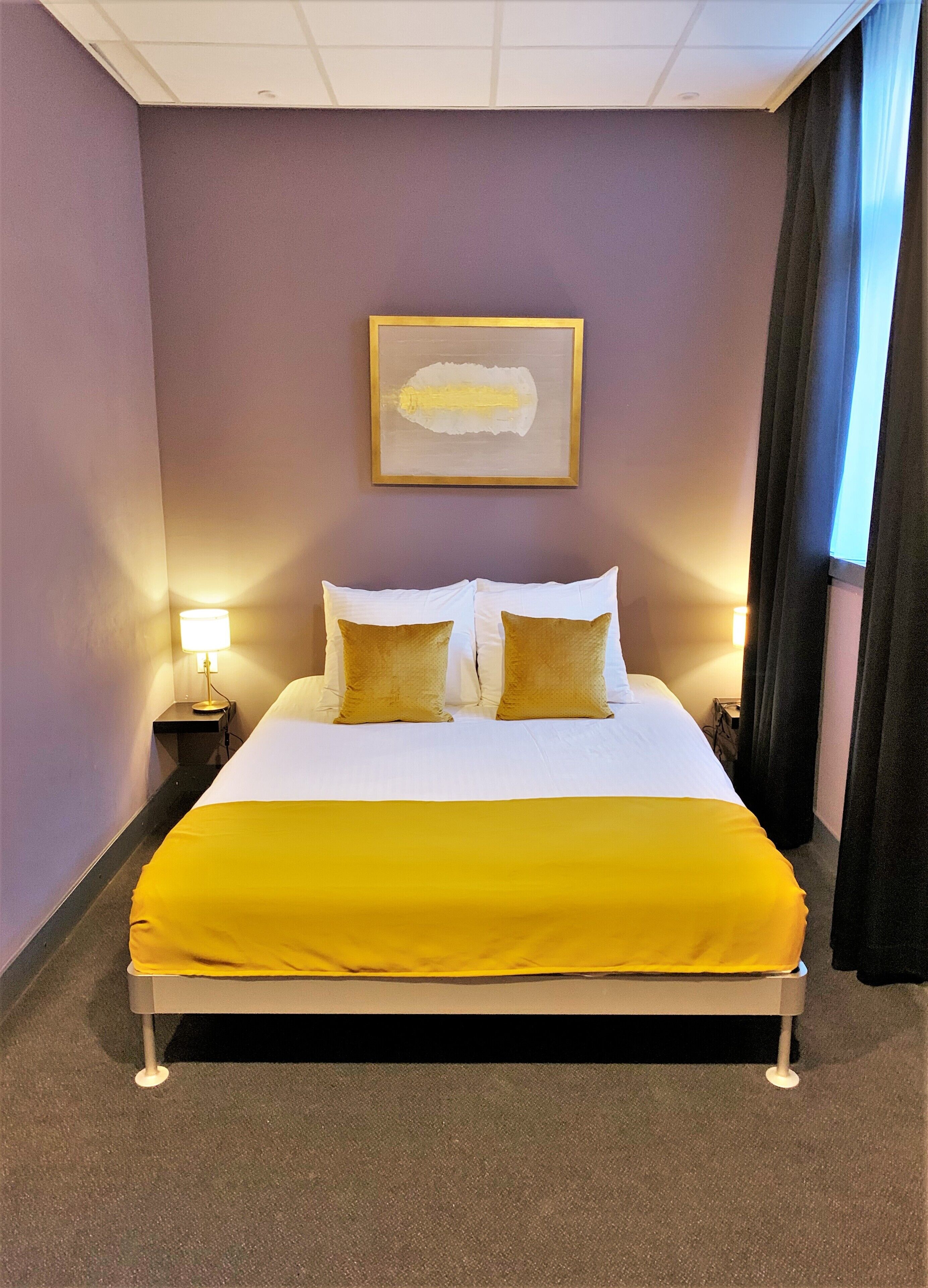 A hotel room with a bed, two bedside tables, a lamp, a framed artwork on the wall, and a window with curtains.