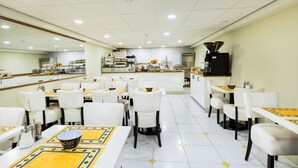 A restaurant with white chairs and tables, a central island with a coffee machine, and a kitchen area in the background. - Hotel De Looier (Amsterdam)