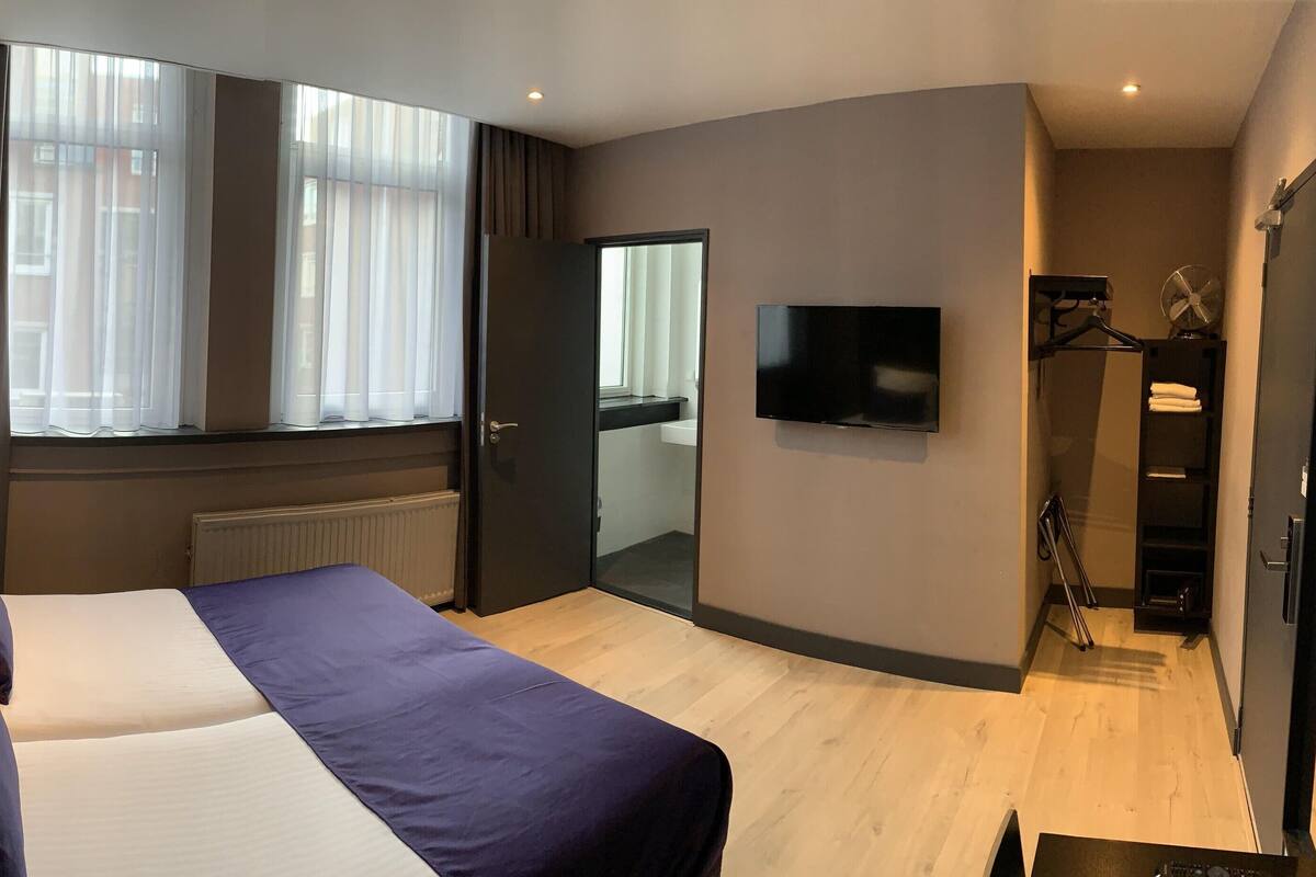 Una habitación de hotel moderna con una cama grande, un televisor de pantalla plana, un vestidor amplio y un ventanal con cortinas translúcidas.