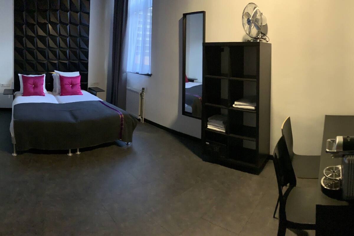 Una habitación de hotel con cama, un escritorio pequeño con silla, una estantería y un espejo.