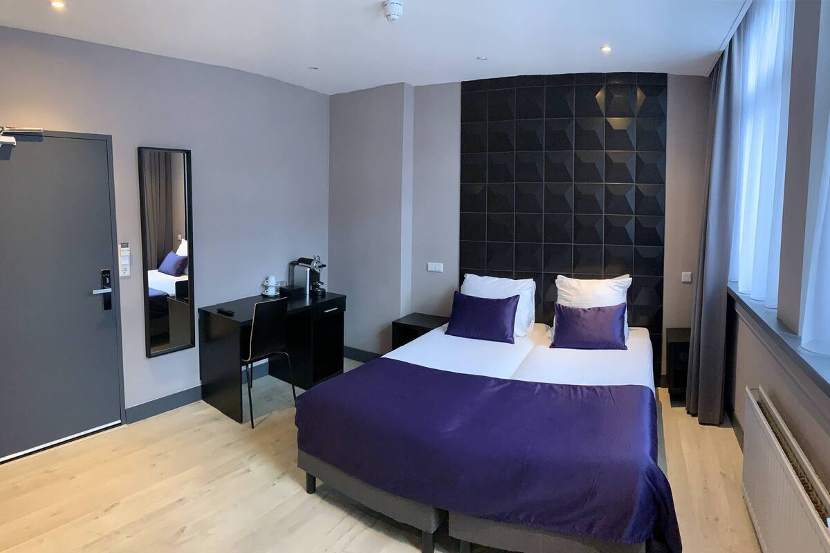 A modern hotel room with a large bed, a desk, a chair, a mirror, and a window with curtains.
