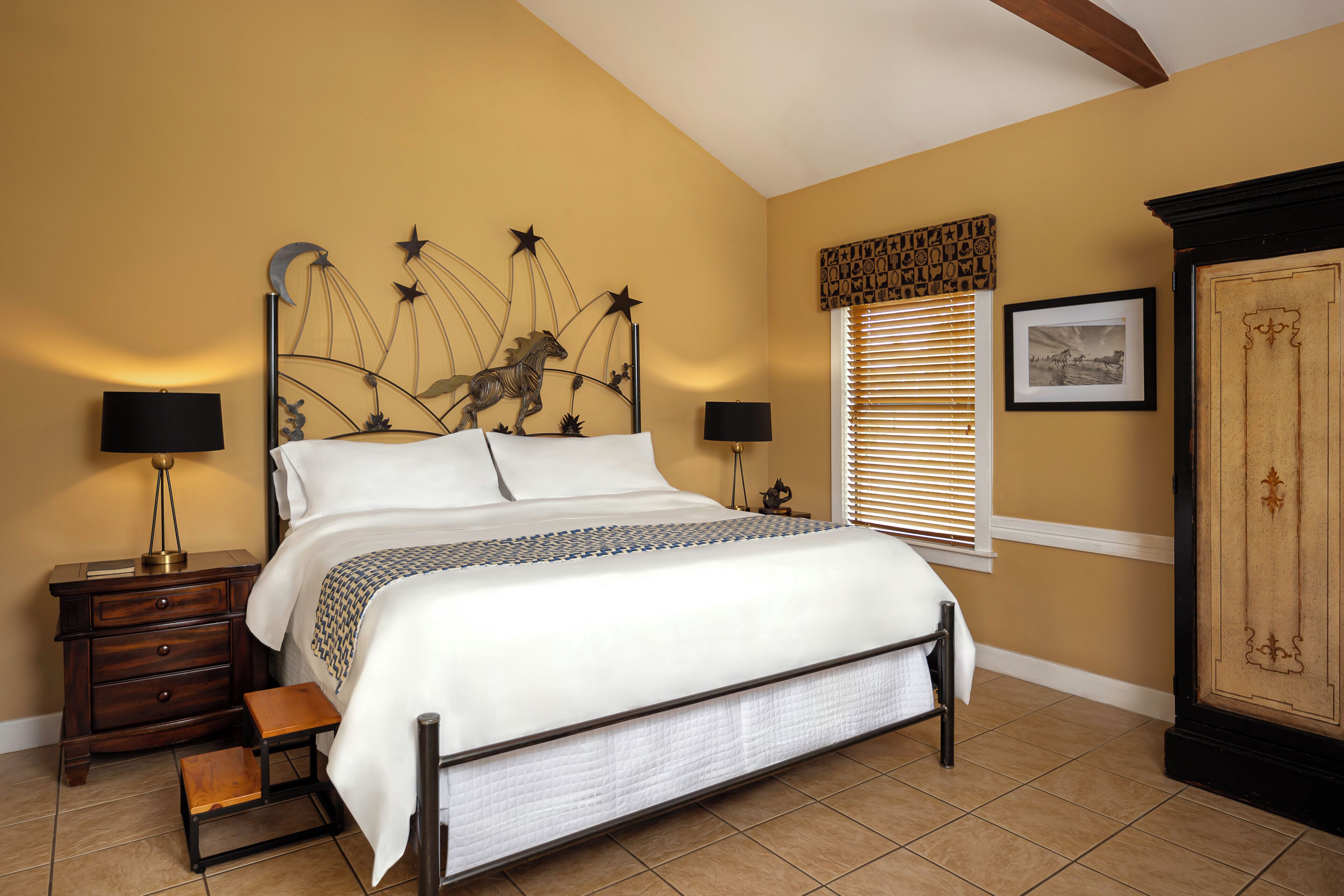 A bedroom with a bed, nightstands, a small table, a cabinet, and a window with blinds.