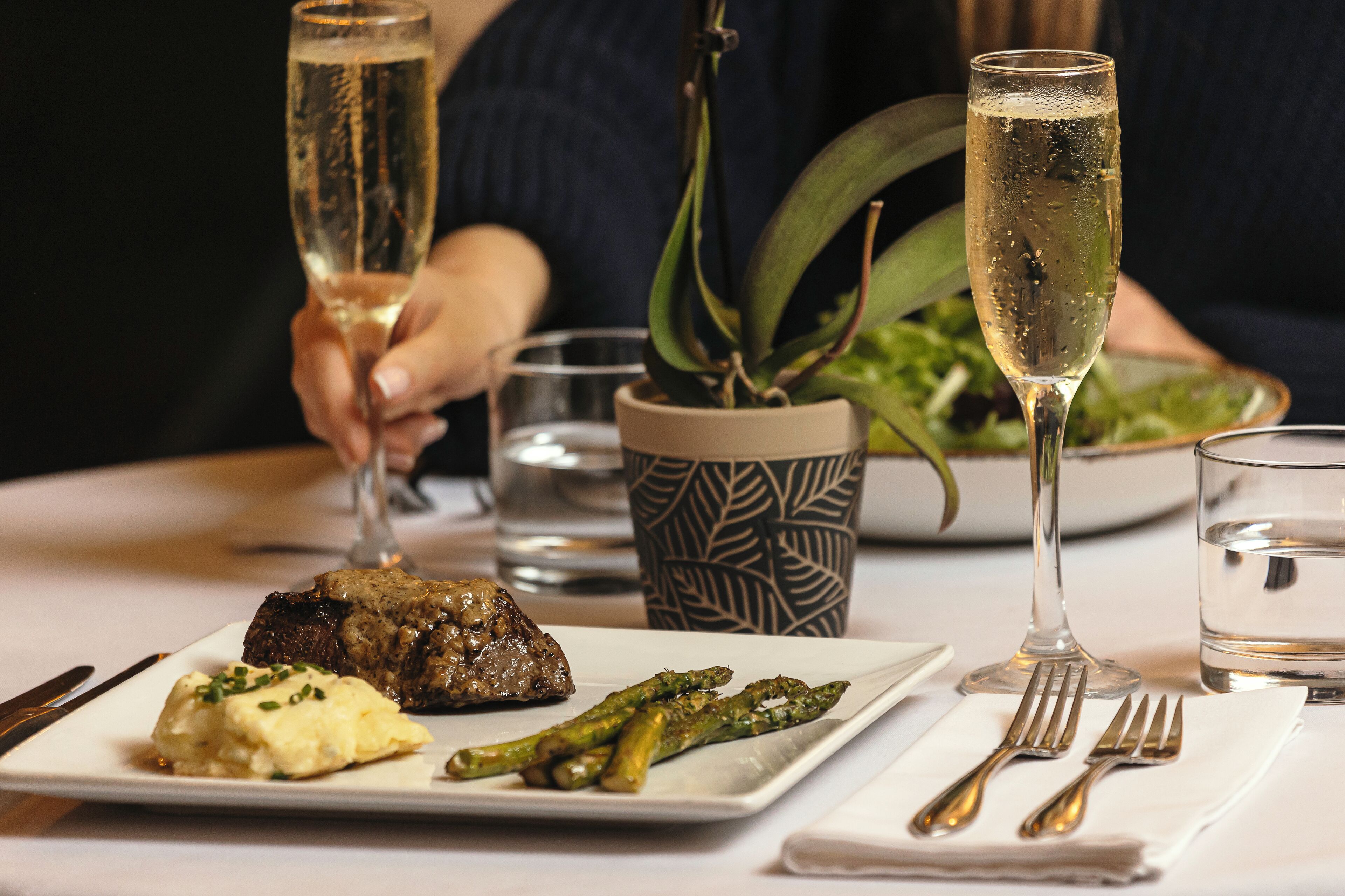 A dining table with a plate of food, a glass of champagne, and a potted plant.