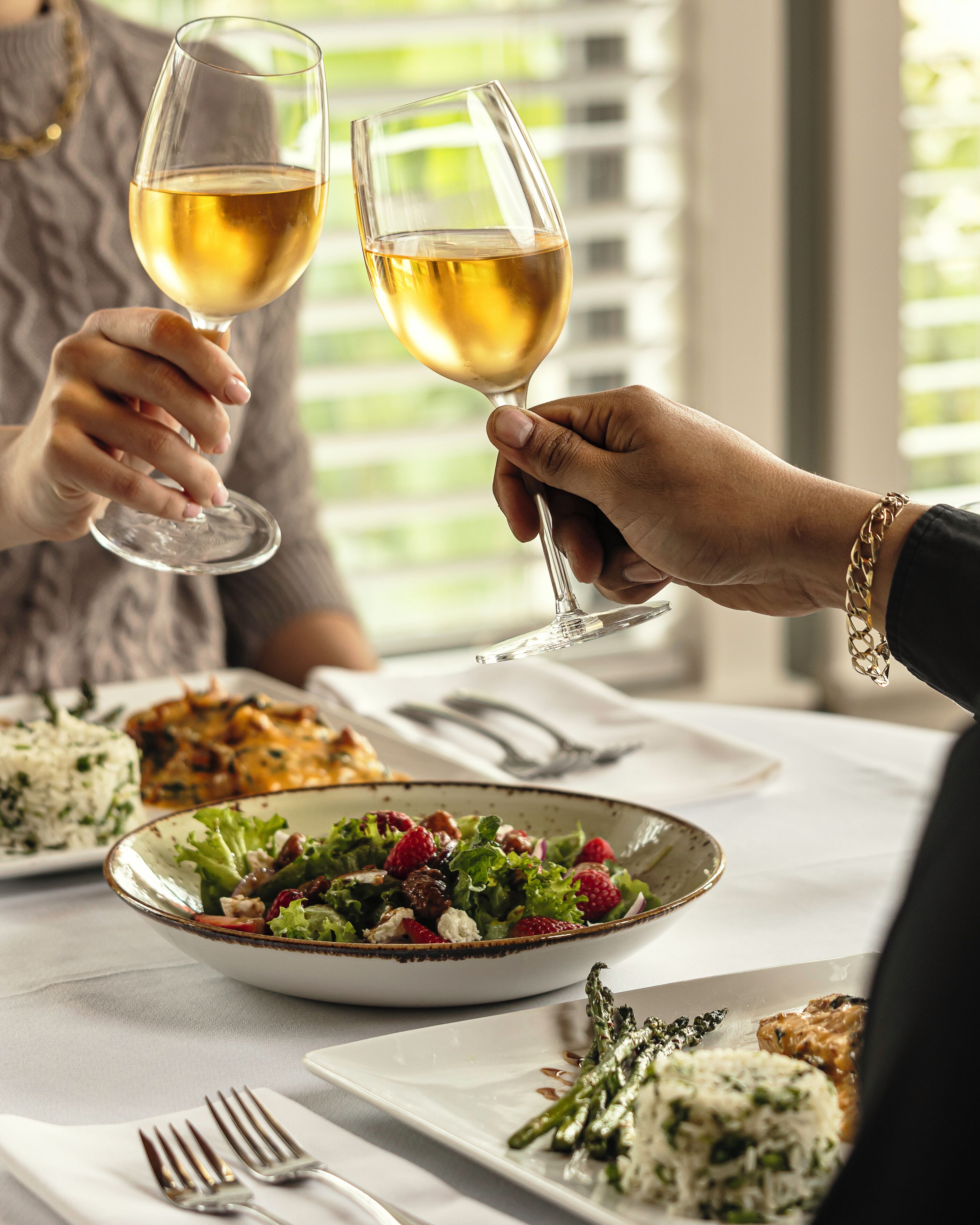 A dining table with plates of food, including a salad and rice dishes, and two glasses of white wine being toasted.