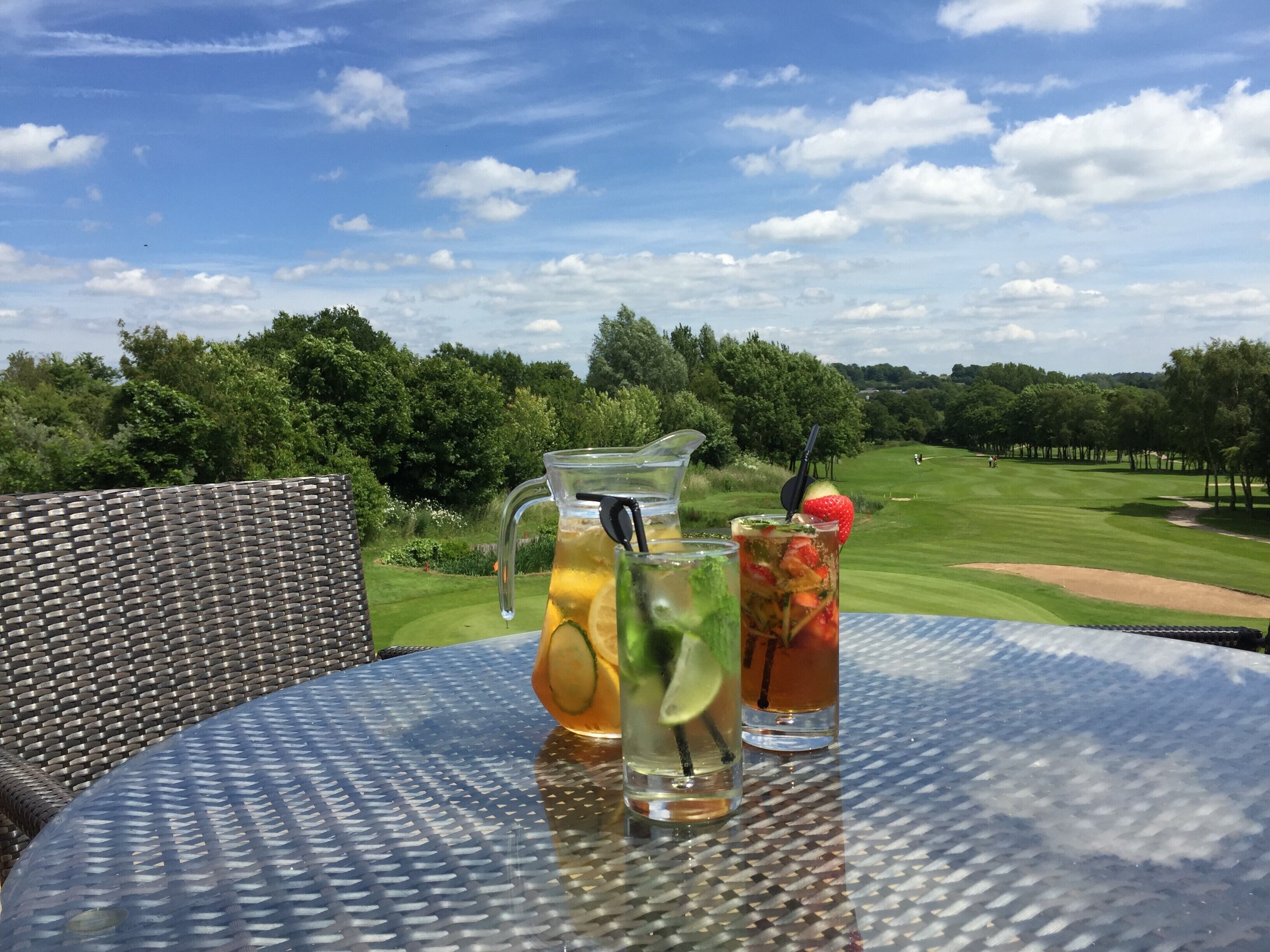 A table with a pitcher and two glasses of iced drinks, a wicker chair, and a view of a golf course.