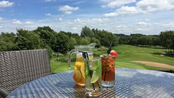 A table with a pitcher and two glasses of iced drinks, a wicker chair, and a view of a golf course.