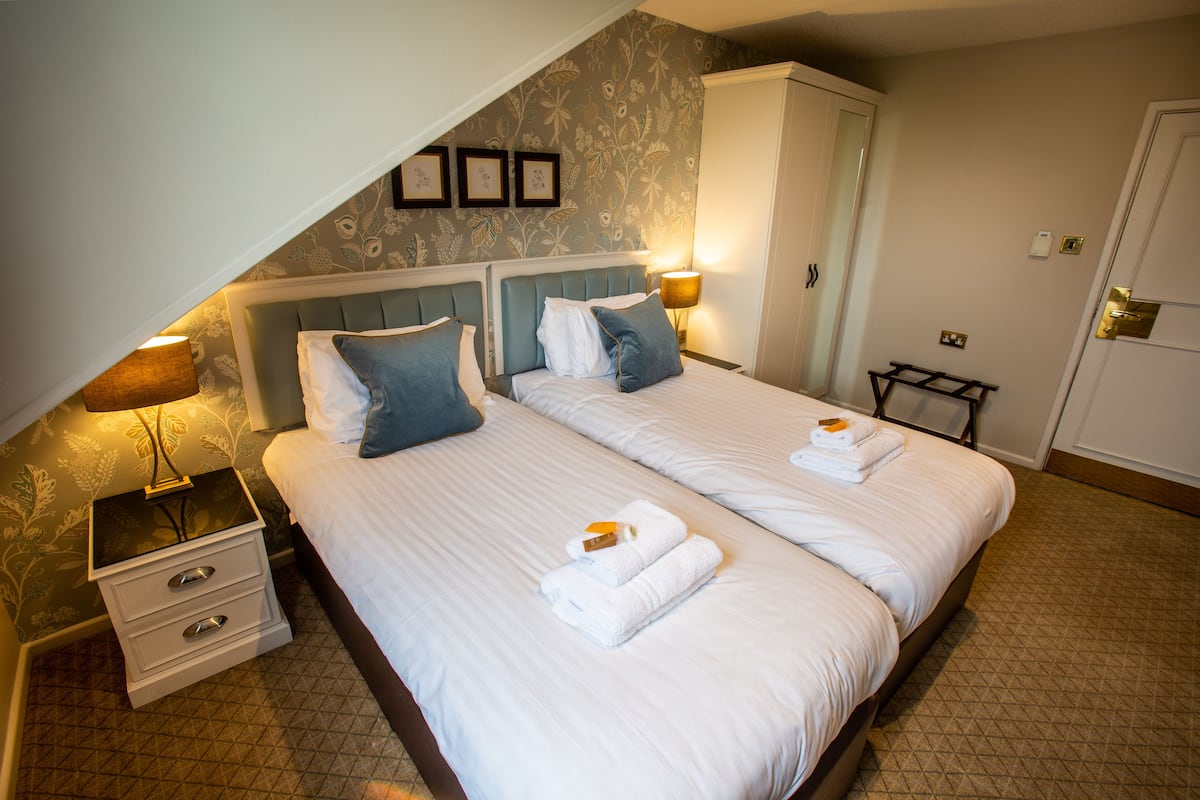 Uma cama de casal com roupa de cama branca e duas almofadas, uma mesa de cabeceira com abajur e um guarda-roupa num quarto de hotel.