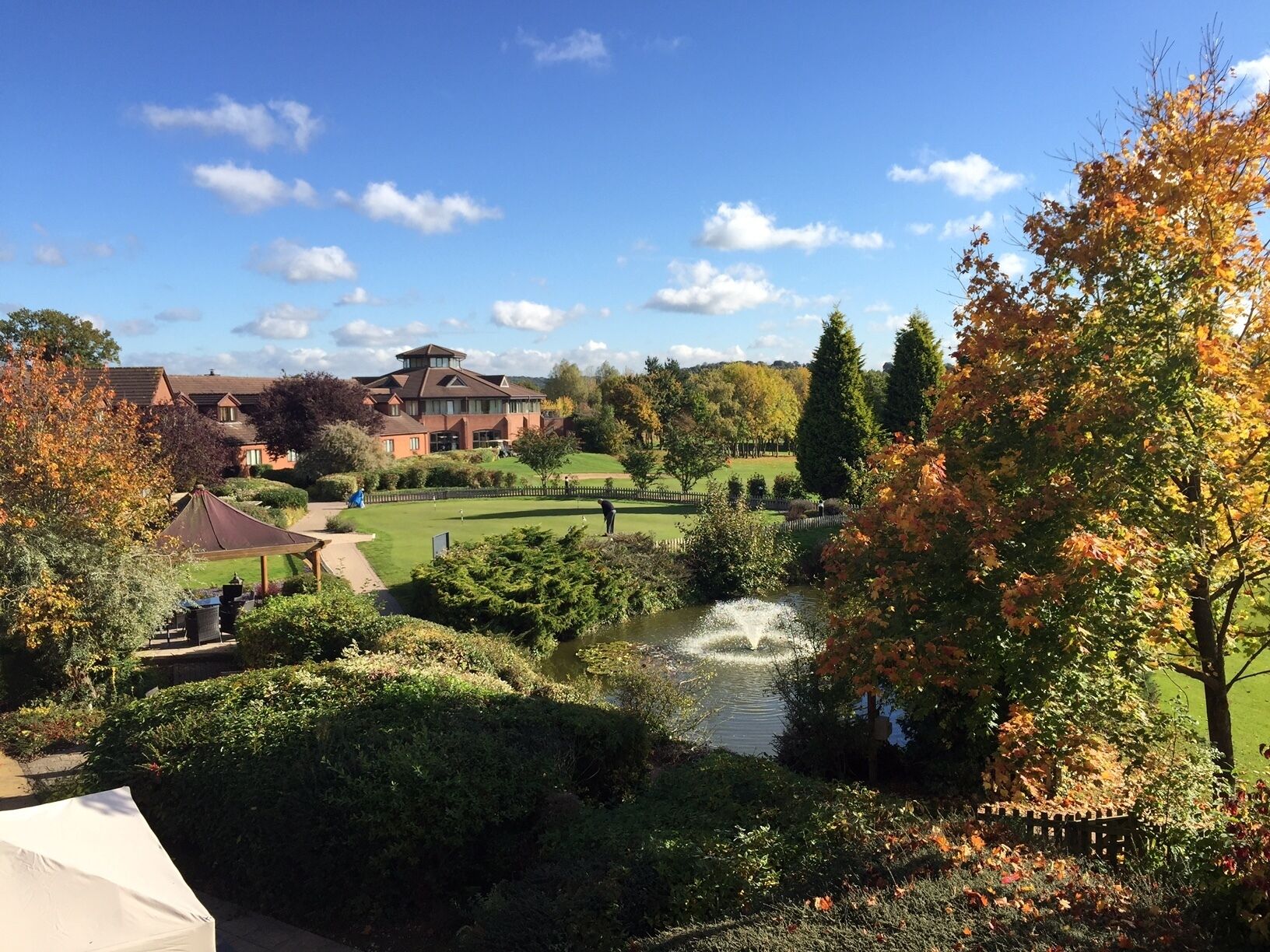 A golf course with a pond, surrounded by autumn foliage and a large building in the background.