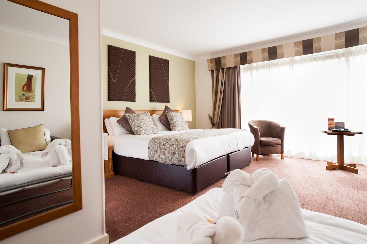 Quarto de hotel com uma cama grande, uma cadeira, uma mesinha e um espelho.
