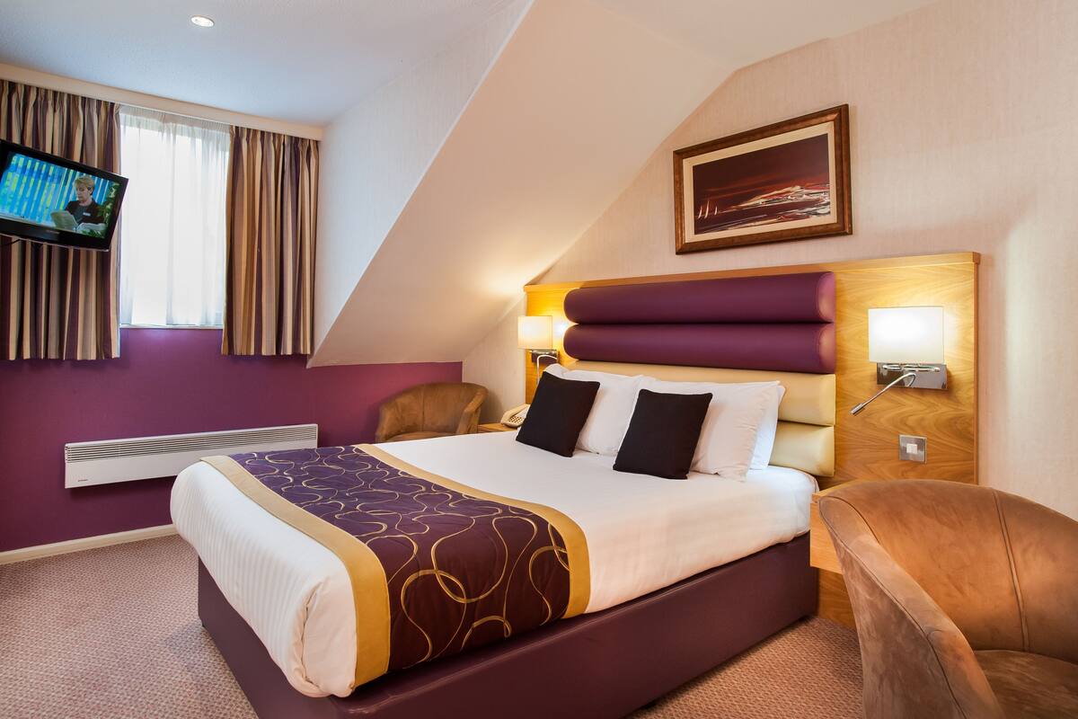 Quarto de hotel moderno com uma cama grande, televisão de tela plana, cabeceira com iluminação embutida, quadro pendurado na parede e aquecedor integrado.