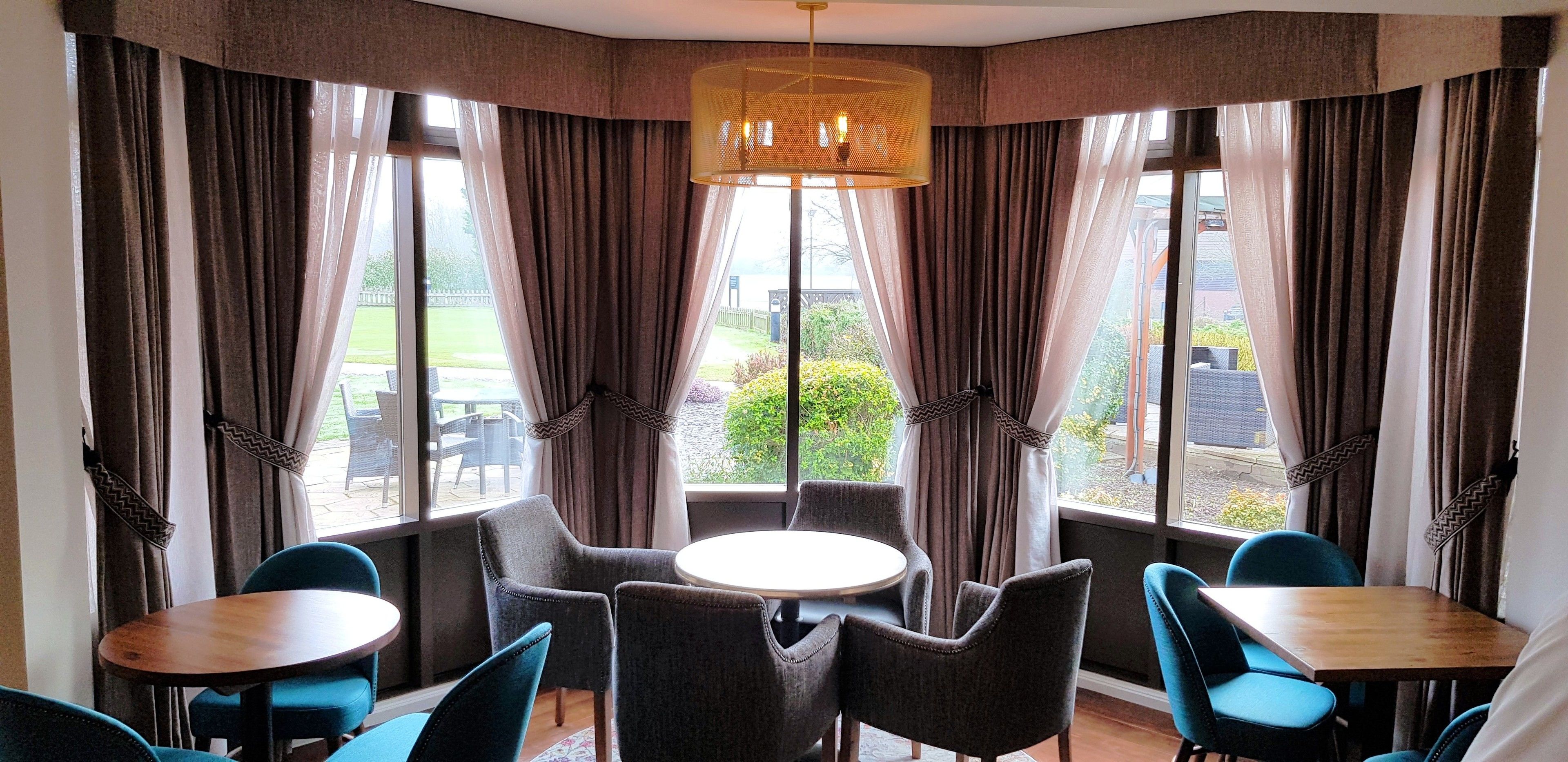 A corner room with a round table and four chairs, large windows with curtains, and a hanging light fixture.