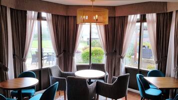 A corner room with a round table and four chairs, large windows with curtains, and a hanging light fixture.