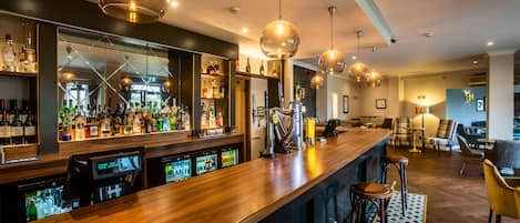 Een bar met een houten toonbank, meerdere hangende lampen en een verscheidenheid aan flessen en glazen.