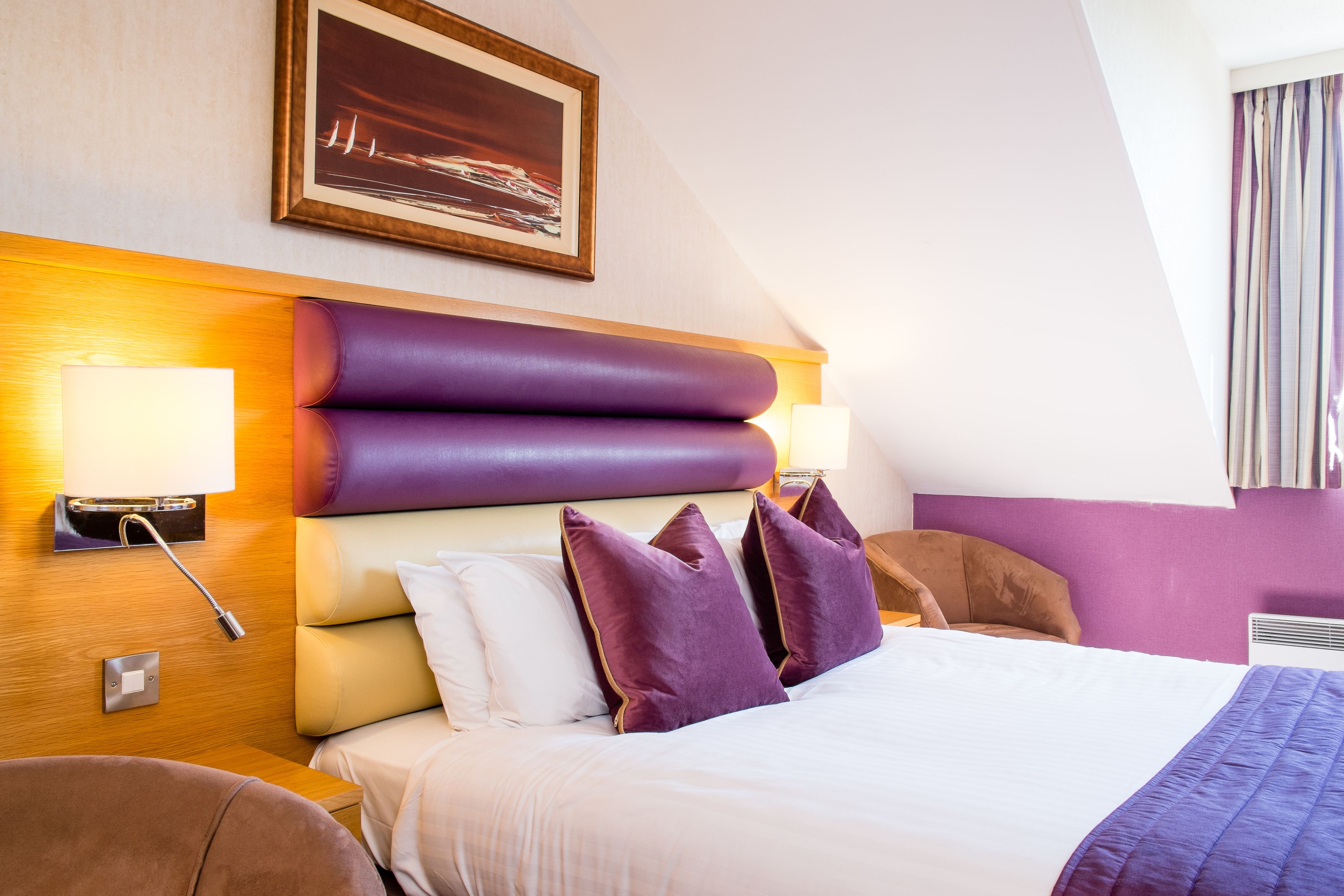 A hotel room with a large bed, purple headboard, and a painting above it.