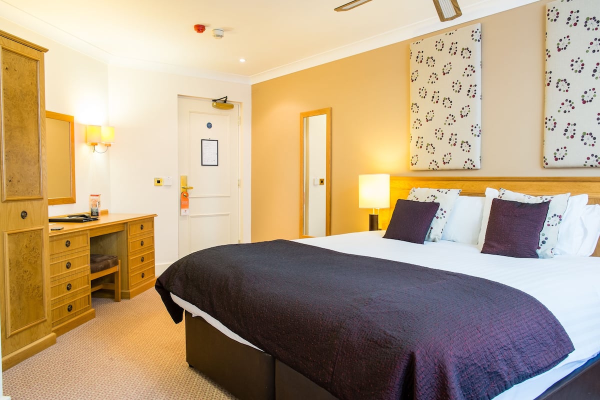 Quarto de hotel com uma cama grande, uma escrivaninha de madeira, uma cadeira, um guarda-roupa e um espelho.
