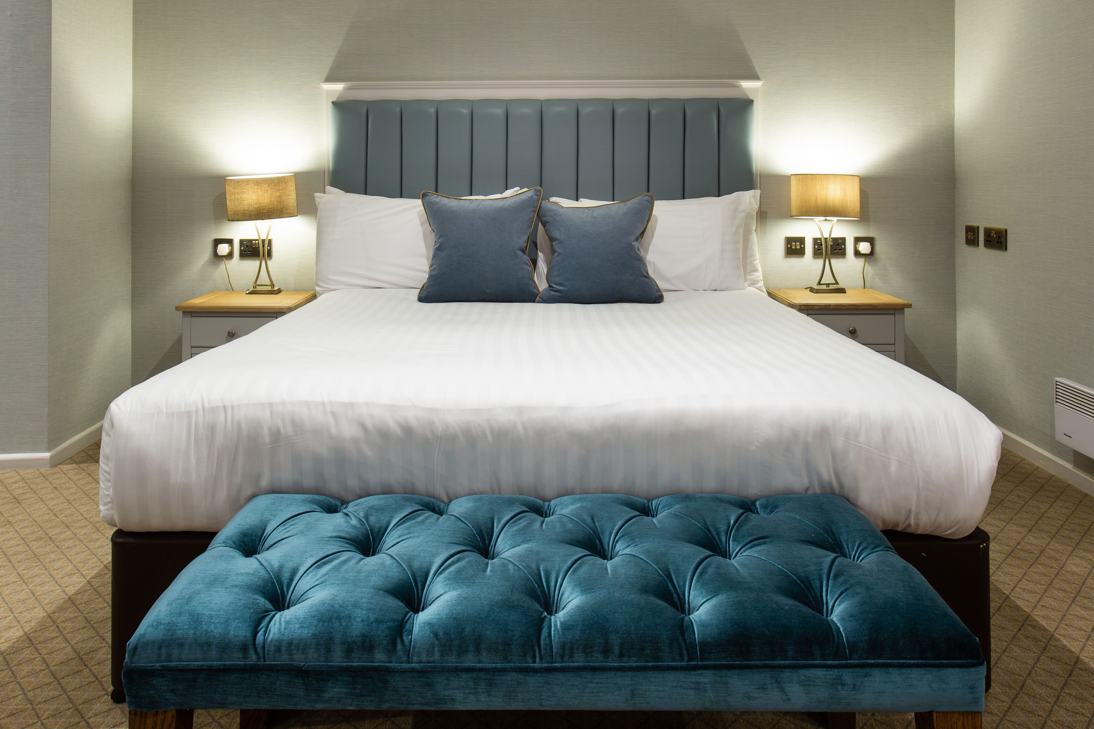 A hotel room with a large bed, two bedside tables with lamps, a bench at the foot of the bed, and a wall-mounted heater.