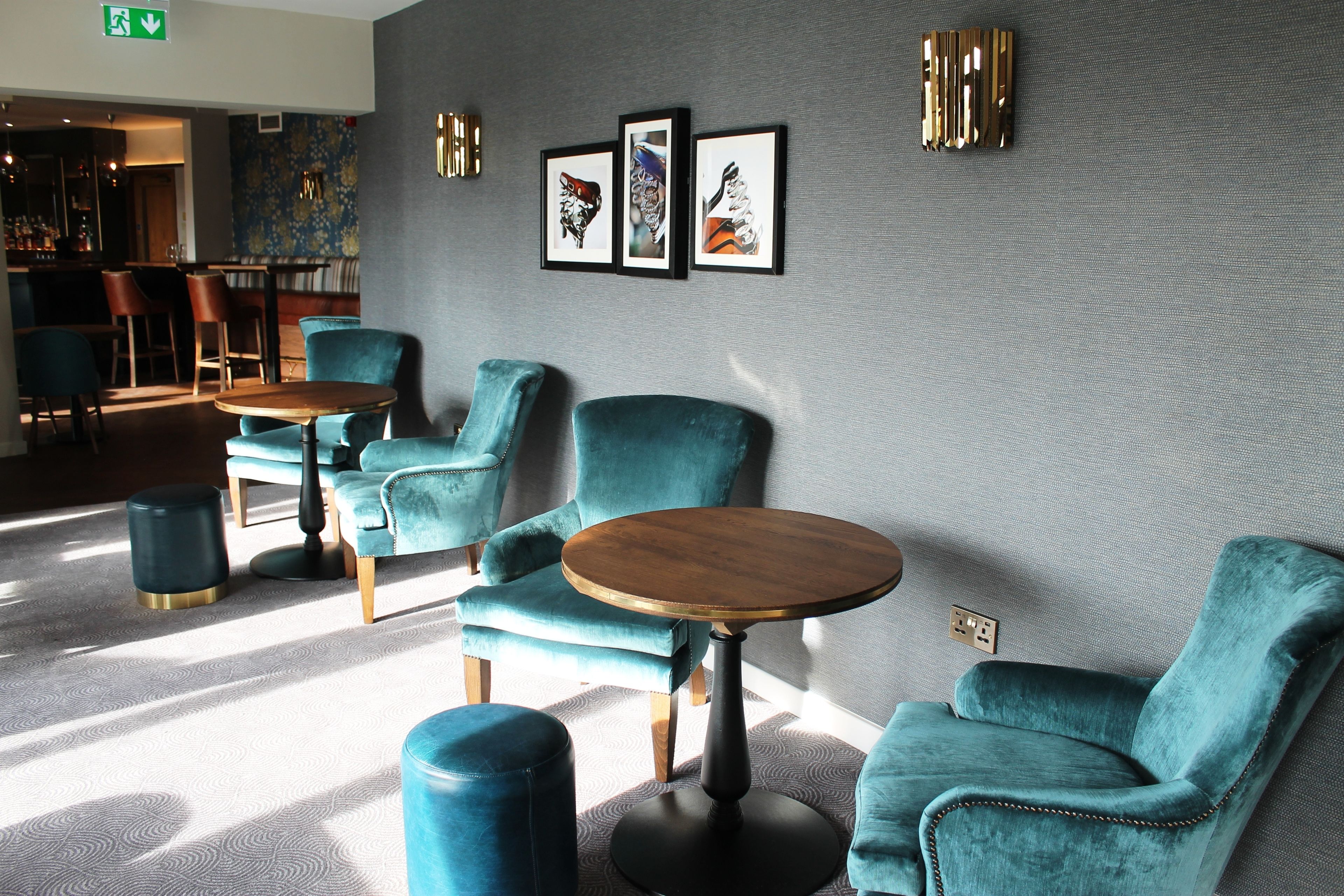 Ein stilvoller Lounge-Bereich mit türkisfarbenen Sesseln und runden Tischen, eine Bar im Hintergrund und gerahmte Kunstwerke an der Wand.