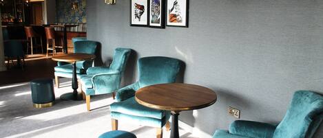 Un área de estar moderna con sillones color turquesa y mesas redondas, una barra al fondo y obras de arte enmarcadas en la pared.
