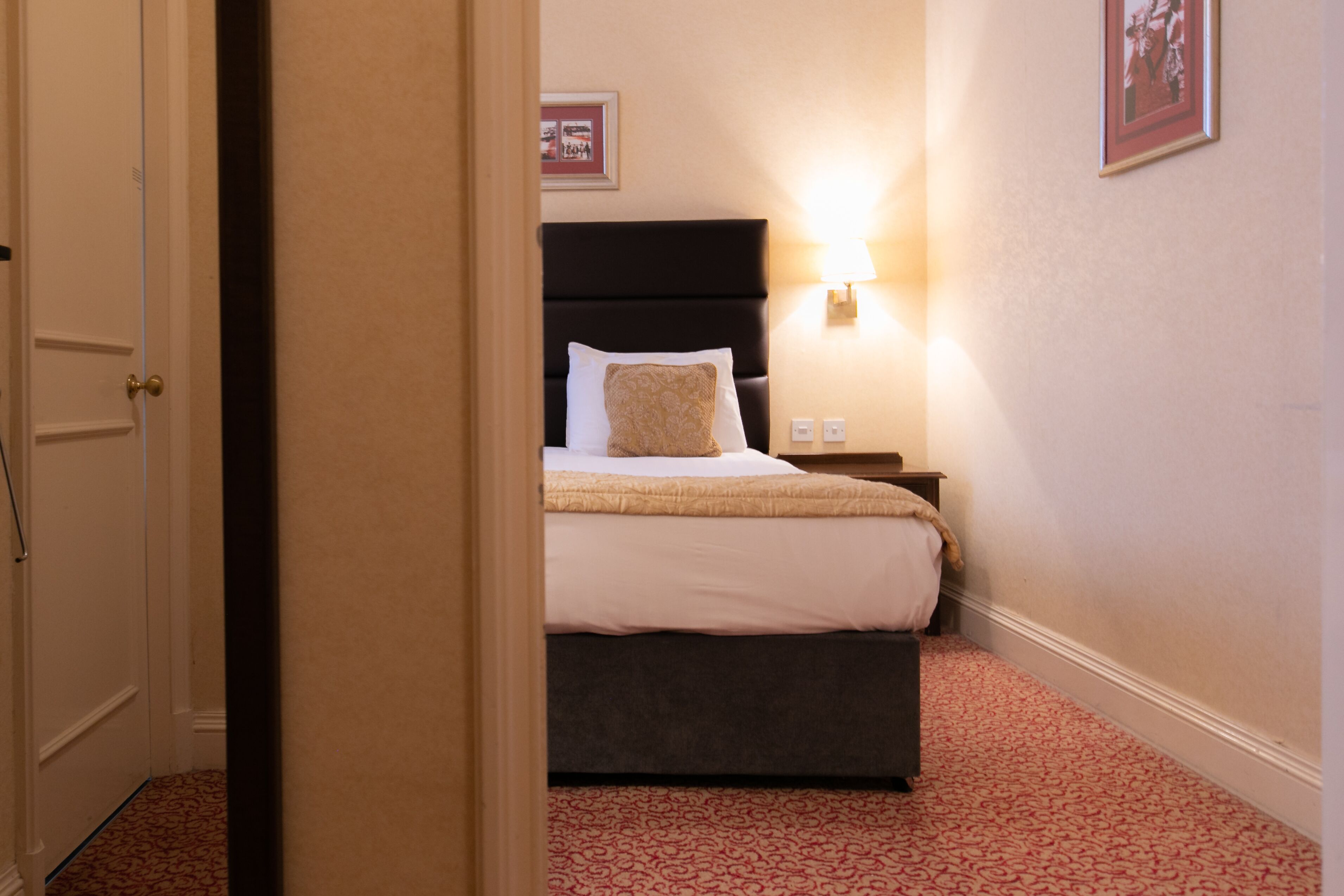 A hotel room with a bed, a nightstand, a wall-mounted lamp, and a framed picture on the wall.