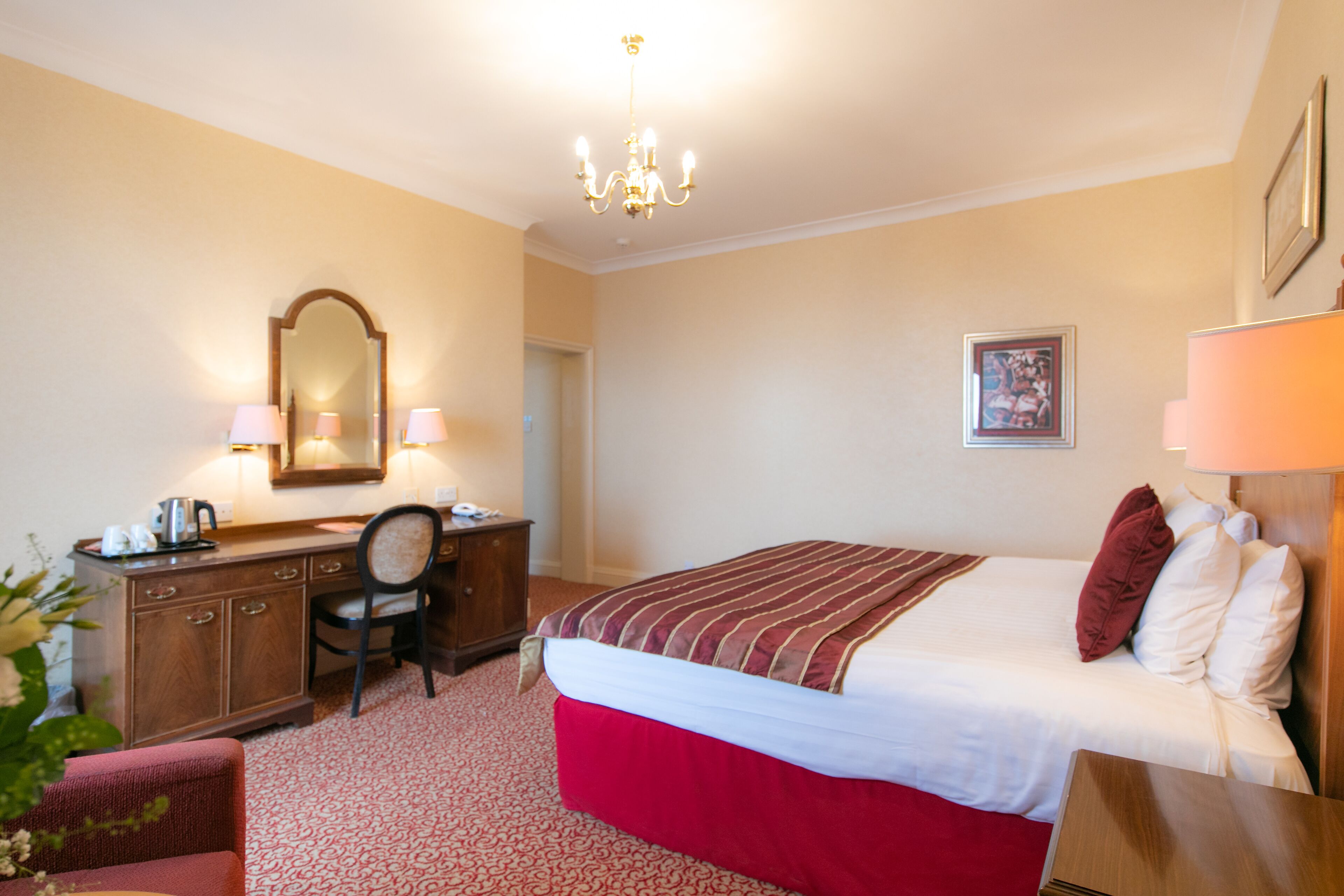 A hotel room with a bed, a desk, a chair, a dresser, a mirror, a chandelier, and a framed picture on the wall.