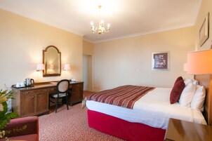 Een hotelkamer met een bed, een bureau, een stoel, een kast, een spiegel, een kroonluchter en een ingelijst kunstwerk aan de muur.