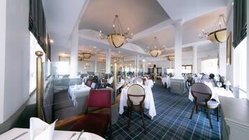 A spacious restaurant with high ceilings, chandeliers, and a checkered floor. Tables are set with white tablecloths and chairs.