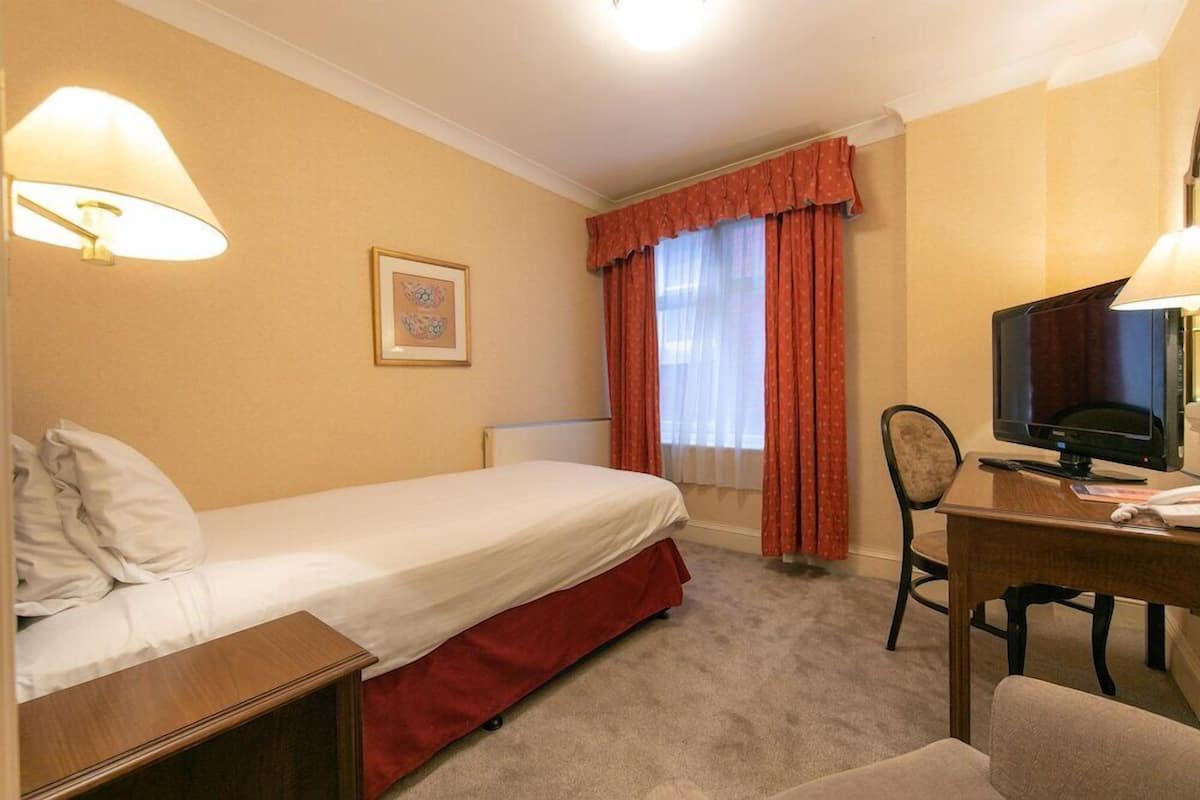 A hotel room with a single bed, a desk with a computer, a chair, and a window with curtains.