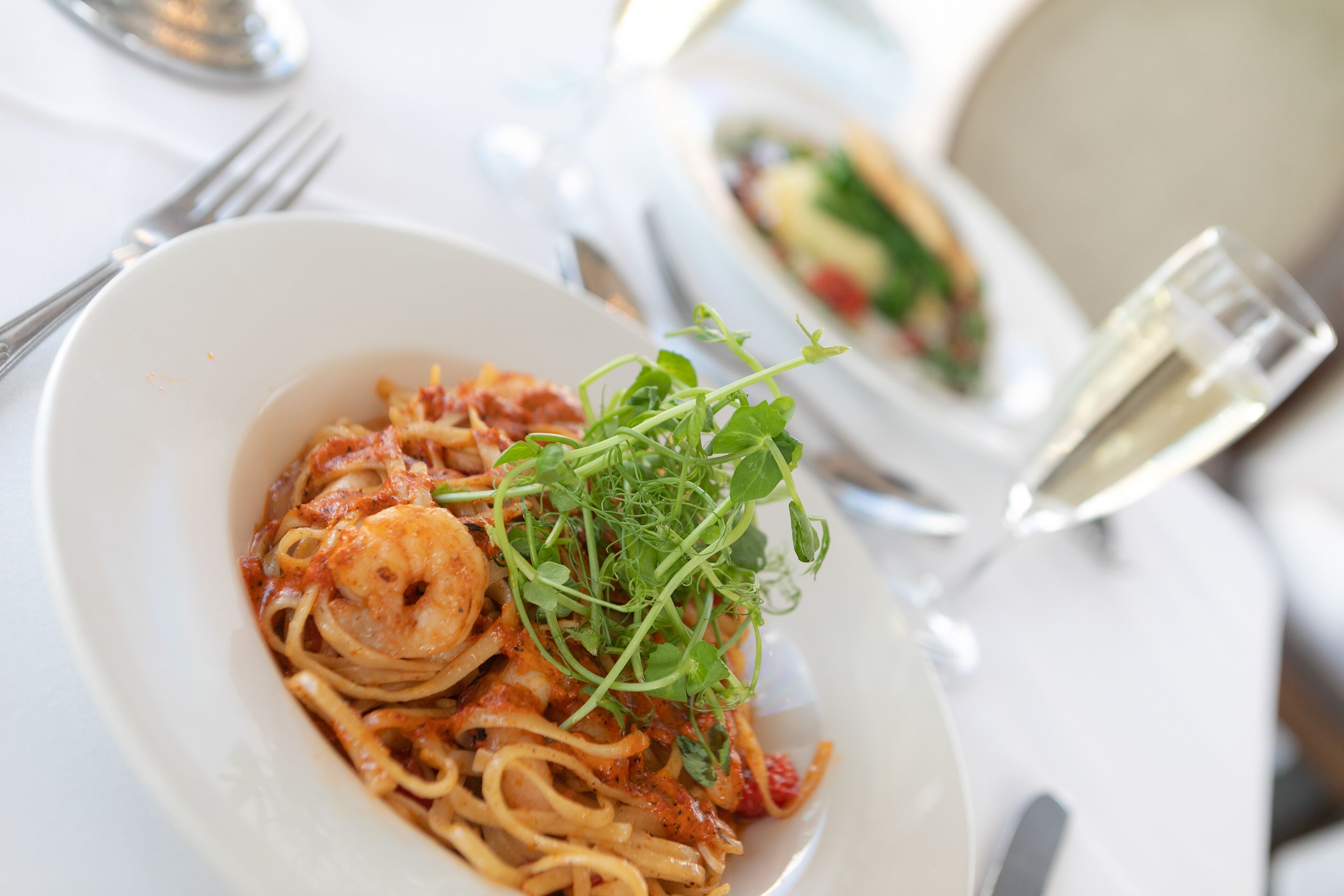 Un plato de pasta con camarones y vegetales verdes, un tenedor y un vaso de agua sobre una mesa.