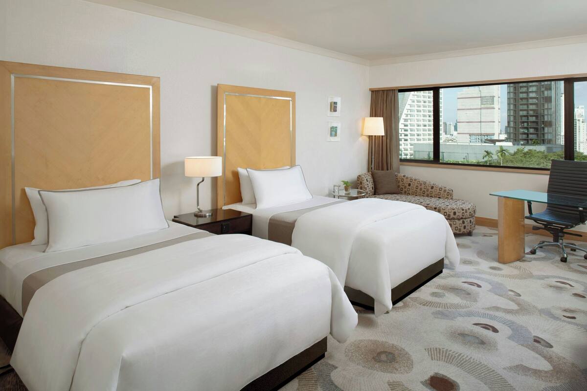 Ein Hotelzimmer mit zwei Betten, einem Schreibtisch, einem Stuhl und einem großen Fenster mit Blick auf die Stadt.