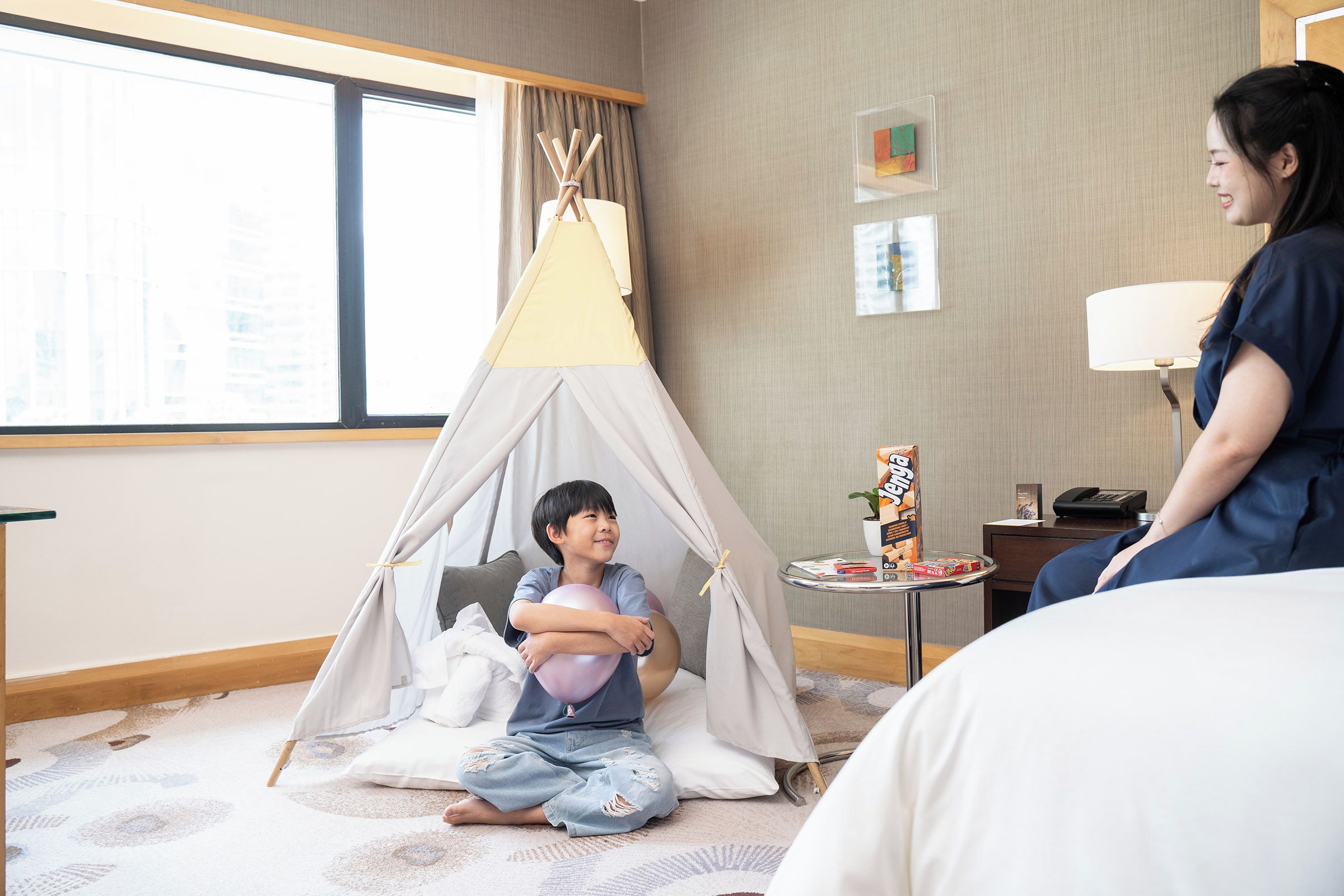 Habitación de hotel con un ventanal grande, una cama, una mesita con una lámpara y un niño jugando dentro de una tienda de campaña.