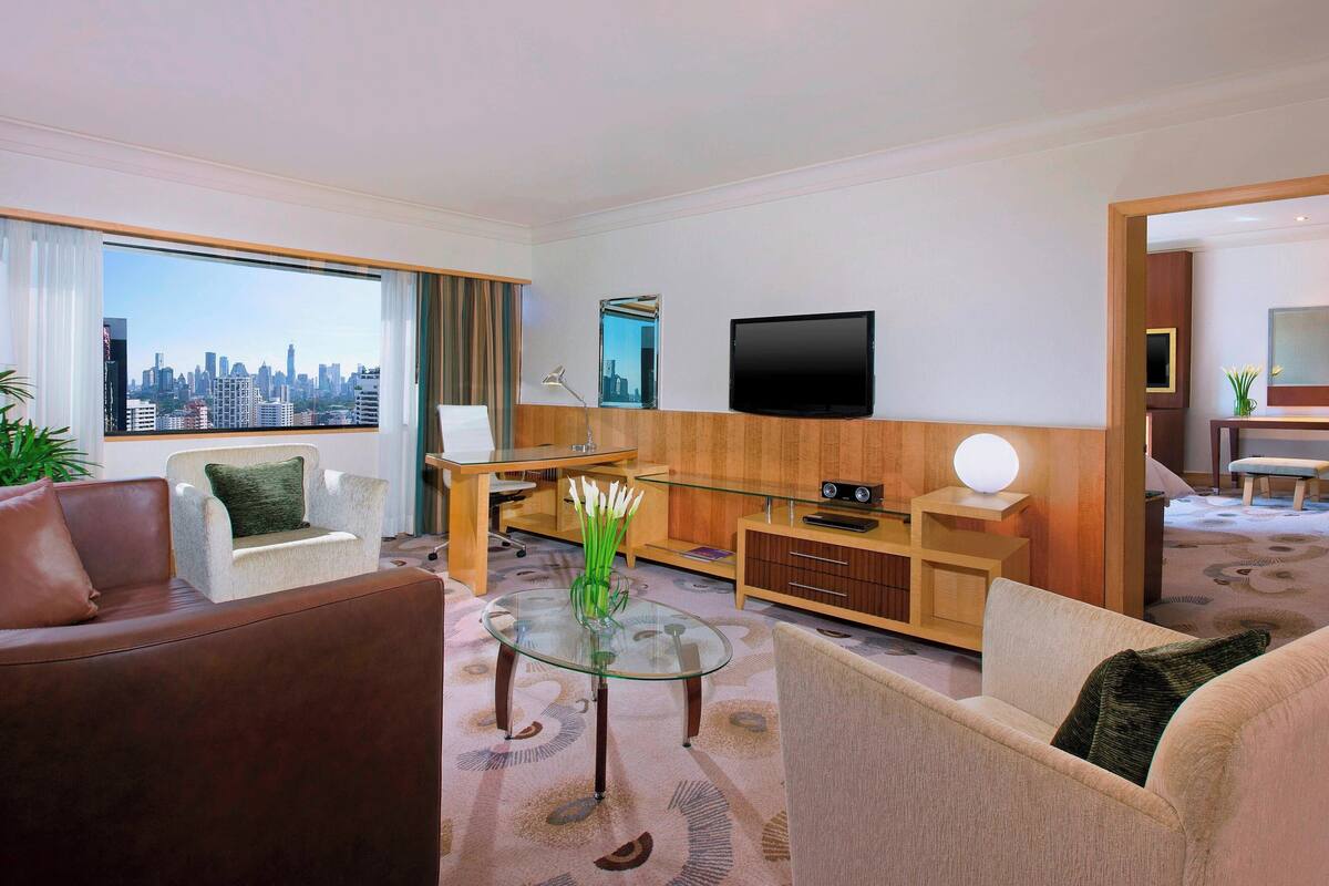 Ein modernes Hotelzimmer mit Blick auf die Stadt, einem Glastisch, Ledersofas, einem Flachbildfernseher und einem Holzkasten.