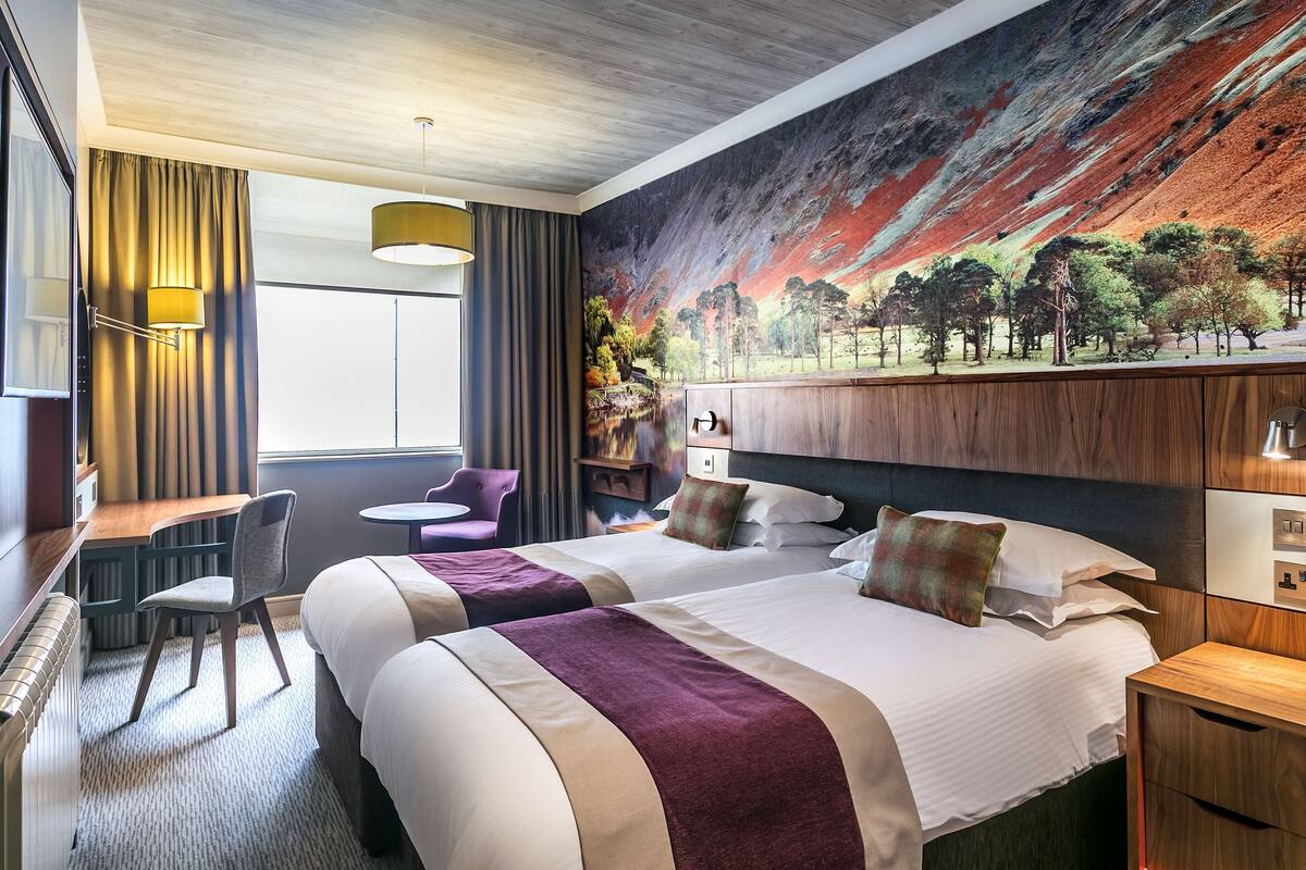 A hotel room with two beds, a desk, a chair, a small table, and a large mural on the wall.