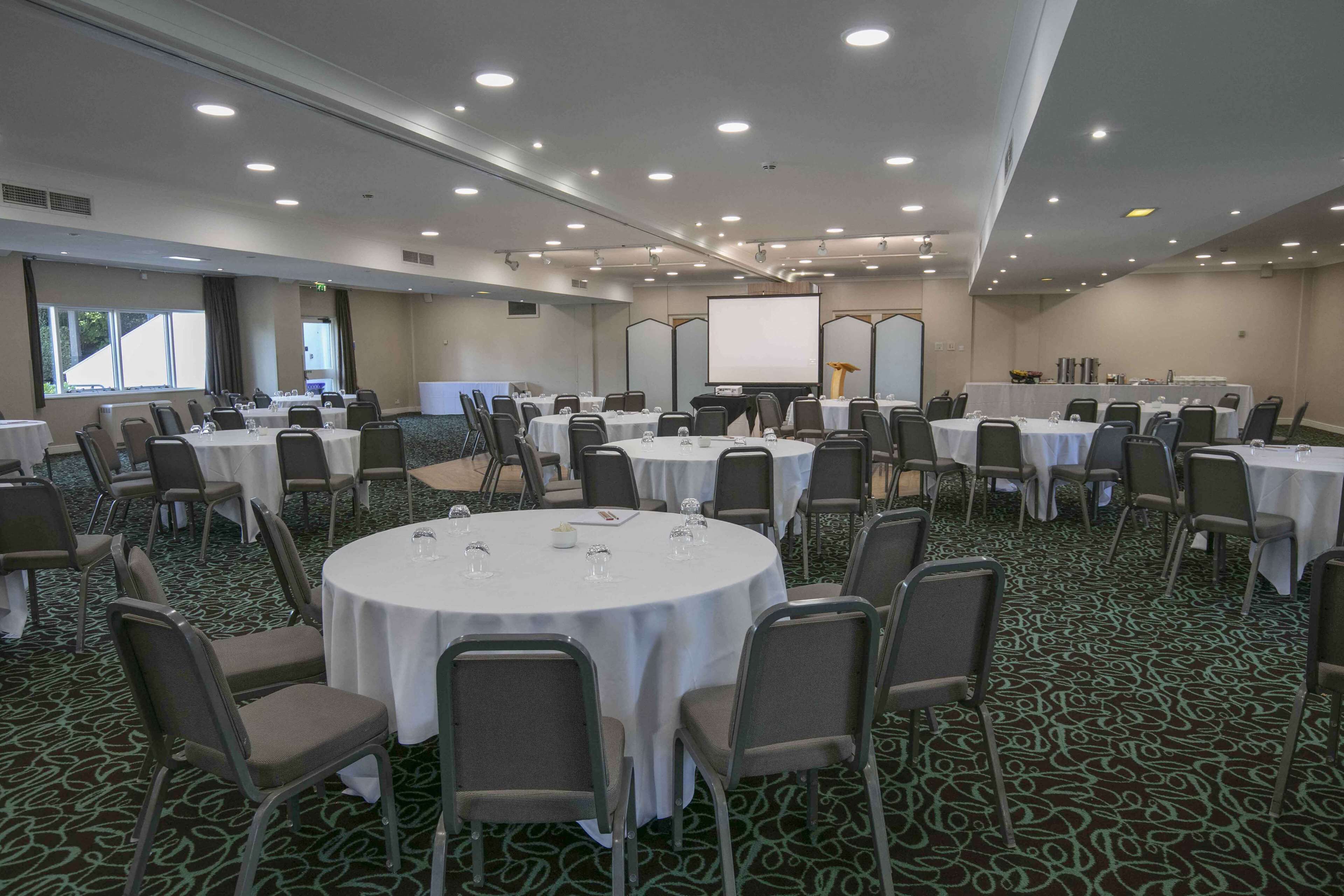 Een vergaderzaal met meerdere ronde tafels, elk omringd door stoelen, en een groot scherm aan de voorkant.