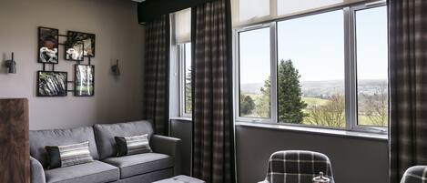 A modern living room with a grey sofa, a matching ottoman, and plaid chairs. There are abstract wall art pieces and a large window with curtains.
