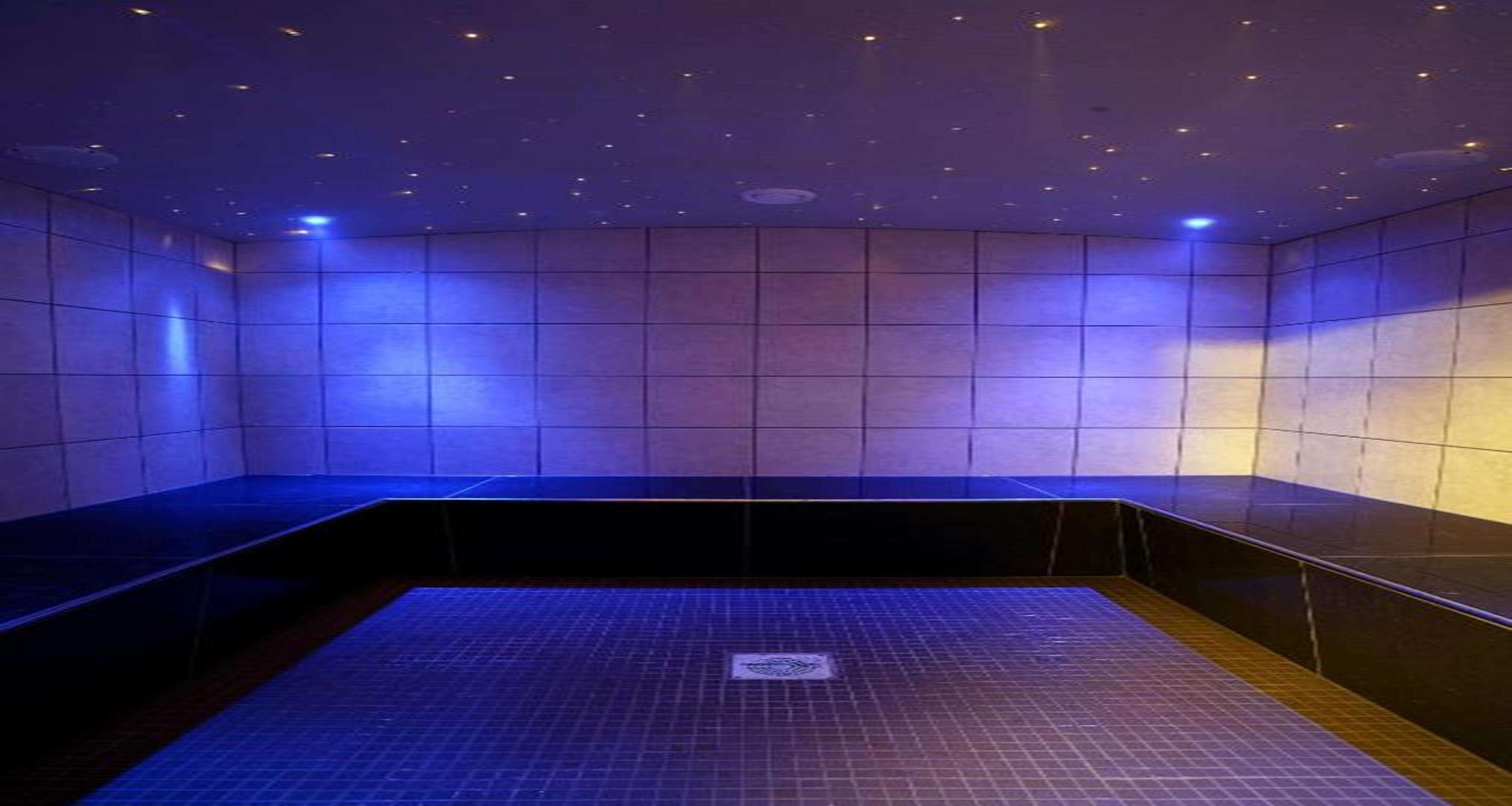 Una habitación moderna, con poca luz, suelo y paredes de azulejos, que incluye un banco y una zona pequeña y cuadrada también de azulejos en el centro.