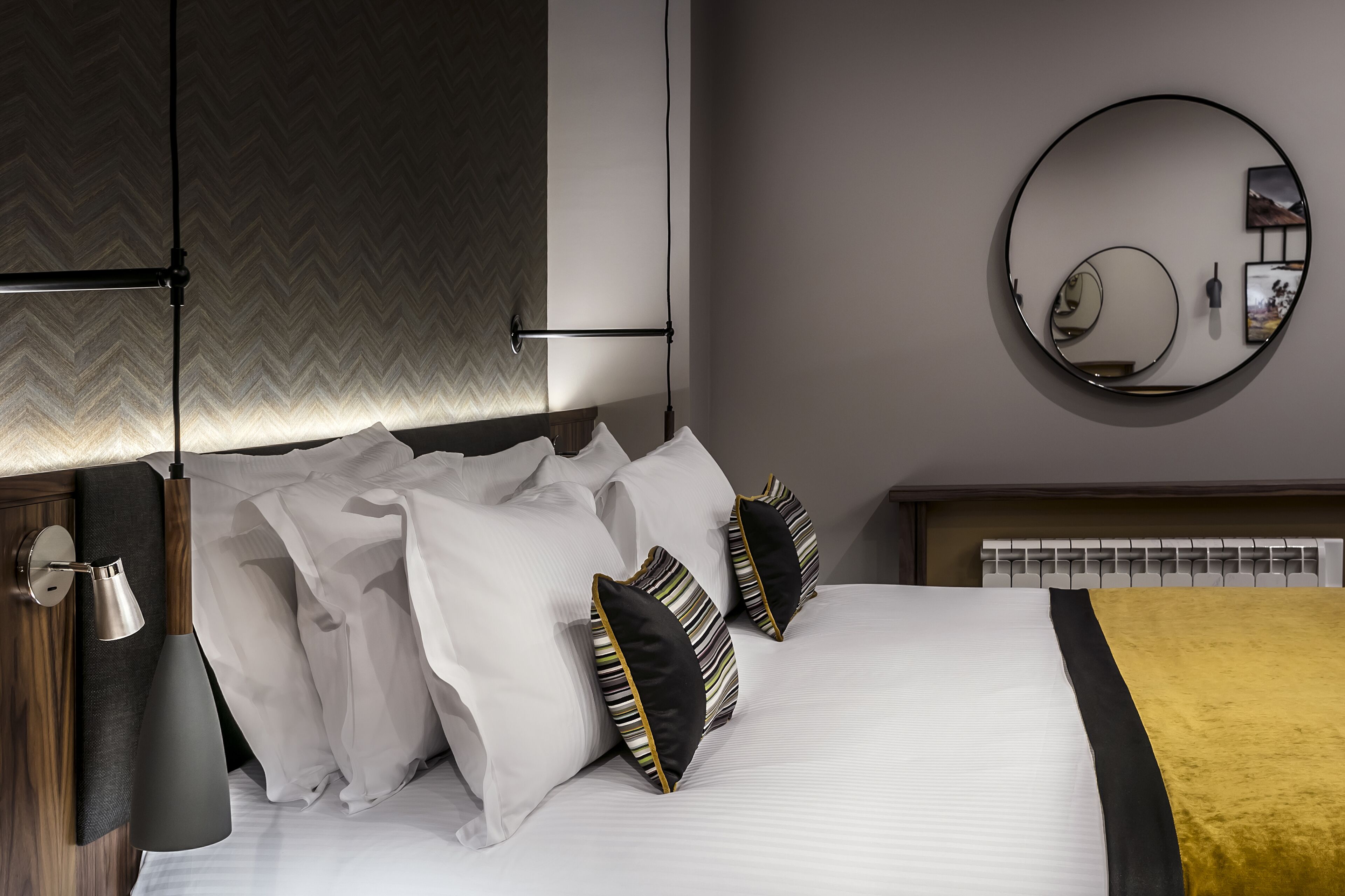A modern bedroom with a large bed, a bedside lamp, a round mirror, and a herringbone-patterned wall.
