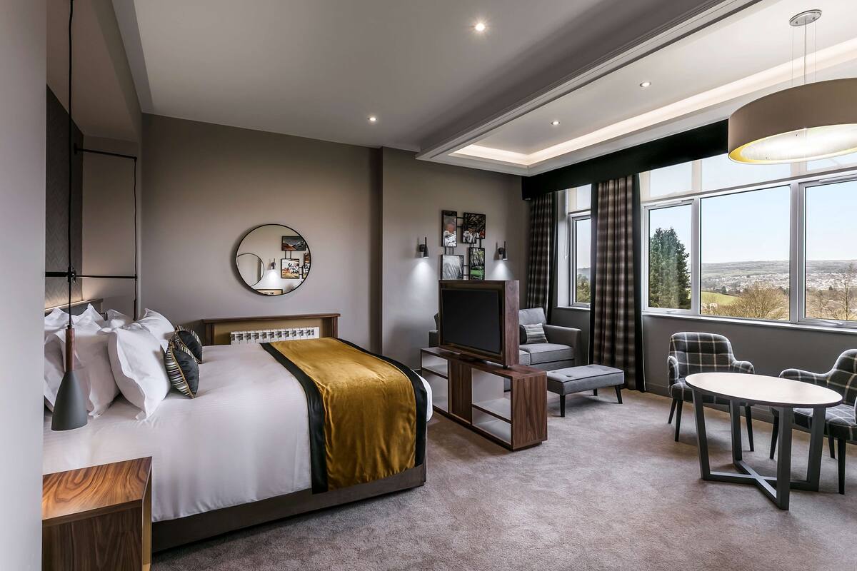 A modern hotel room with a large bed, a TV, a dining area, and a view of the city.